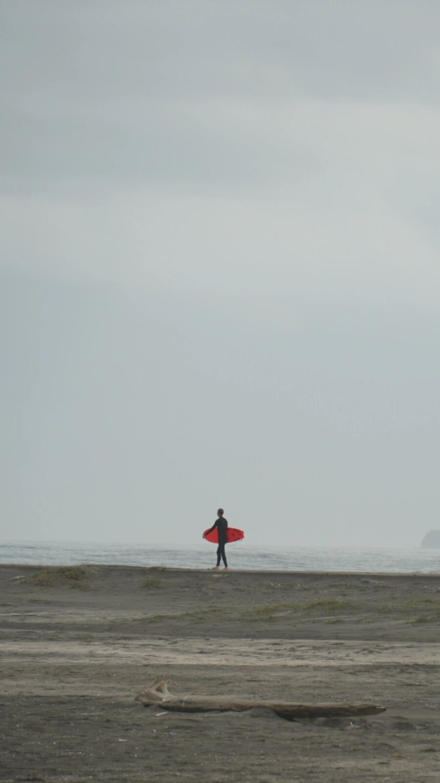 Lone surfer standing on dark sand holding a bright red surfboard, the entire grey Pacific sky behind, driftwood in the foreground