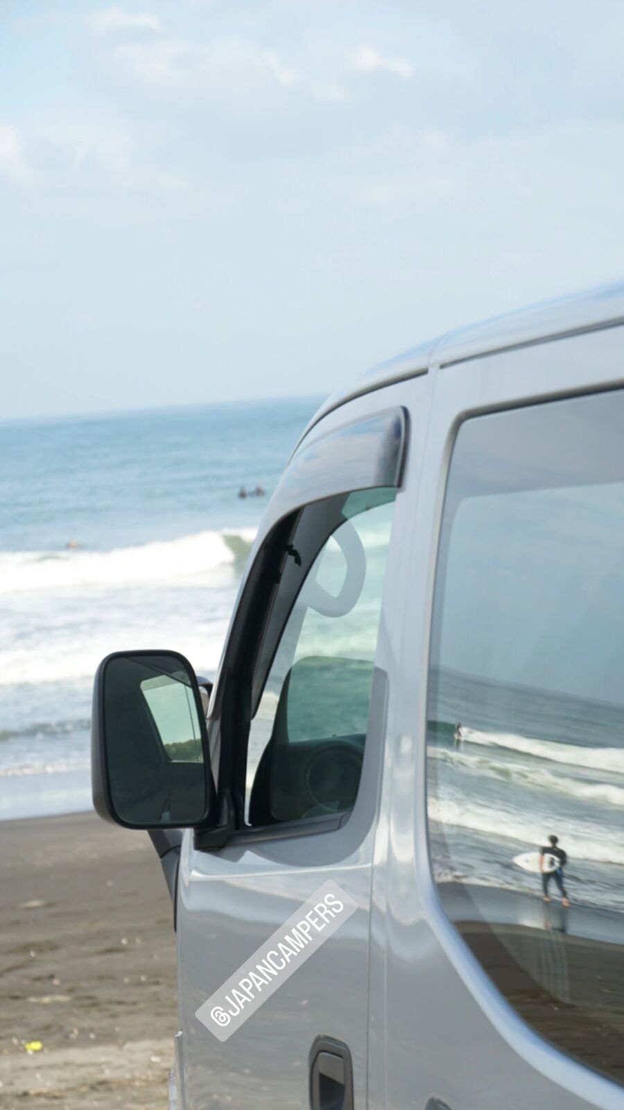 Campervan side mirror reflecting a surfer walking on the beach with the Pacific behind, JapanCampers rental van at Kujukuri
