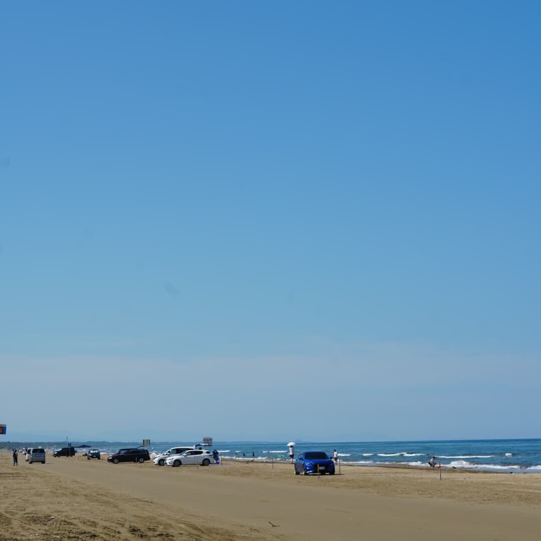 Cars parked across the wide sand beach with swimmers in the shallows, mountains fading into summer haze, the full Chirihama Daytona Beach atmosphere