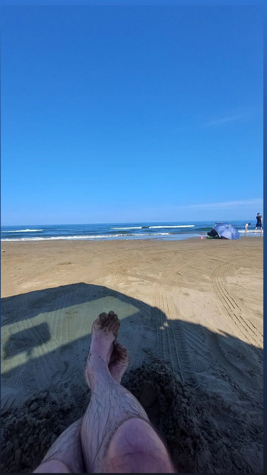 POV from the van's open door: bare feet in the sand, van shadow stretching toward the sea, the beach and distant visitors in the summer heat