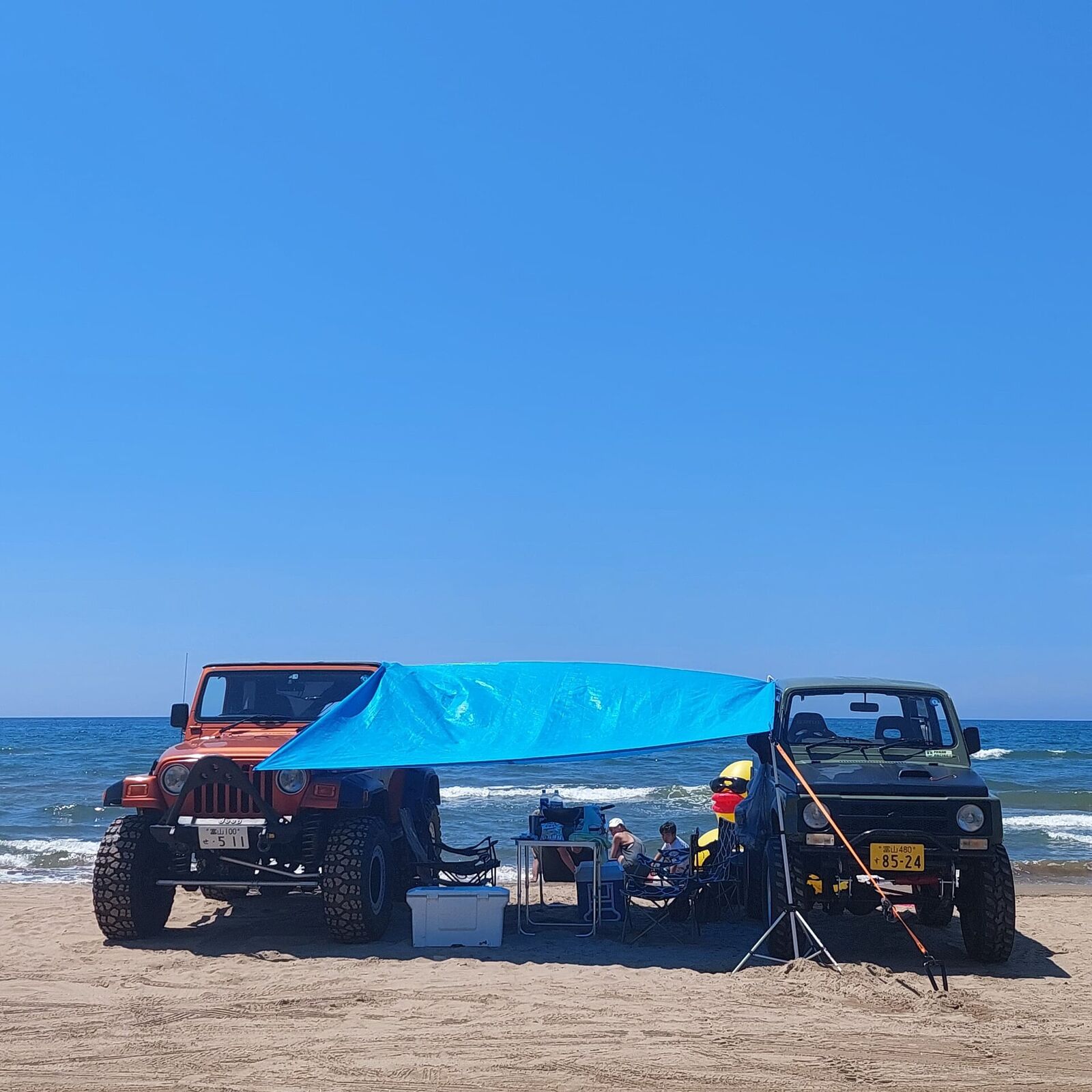A red Jeep Wrangler and green Suzuki Jimny parked nose to nose on the beach, blue tarp stretched between them for shade, camping chairs and cooler box underneath, fishing rod propped up