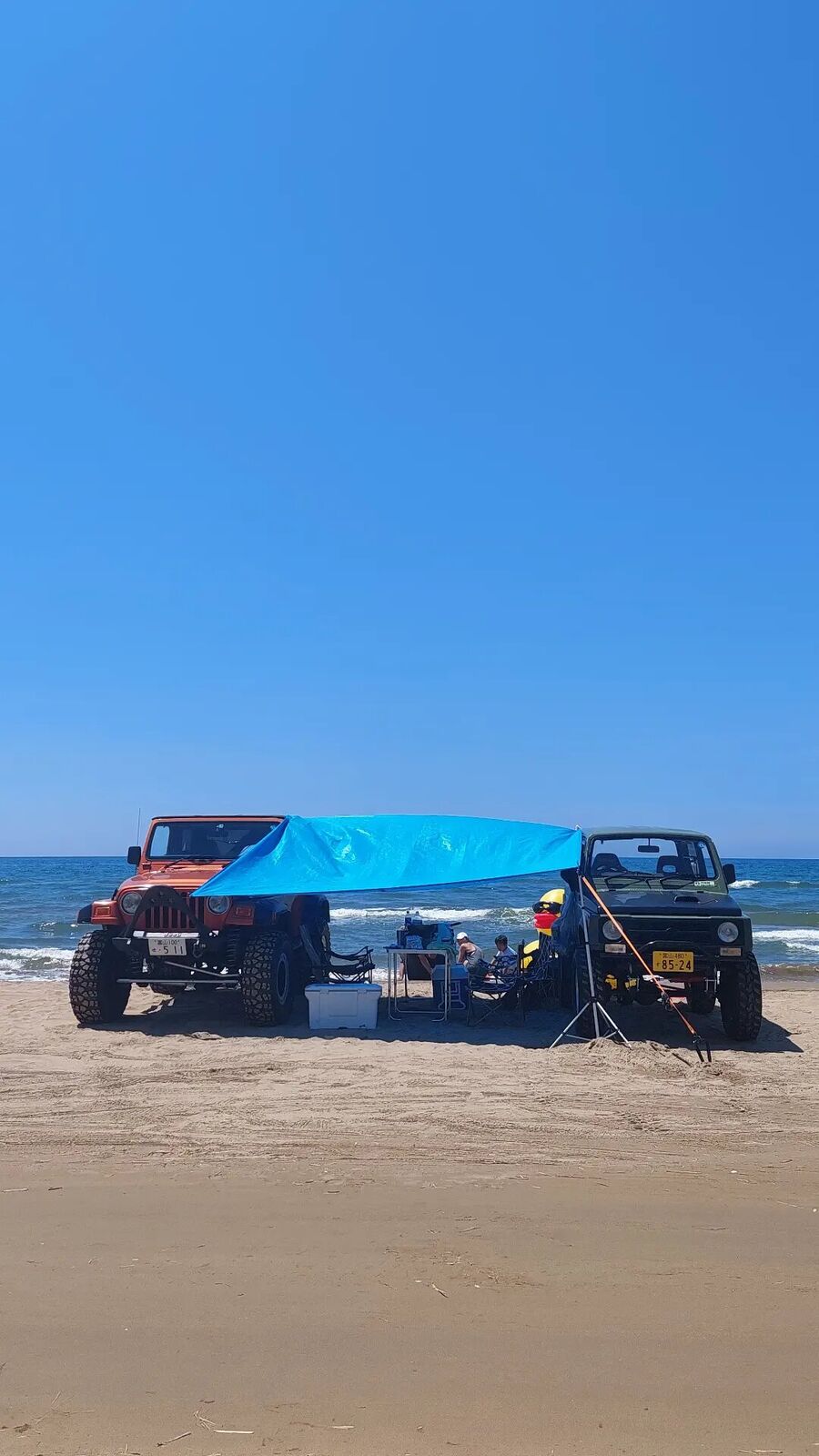 The Jeep and Jimny beach camp from a wider angle, showing the improvised blue tarp shelter with the Sea of Japan right behind
