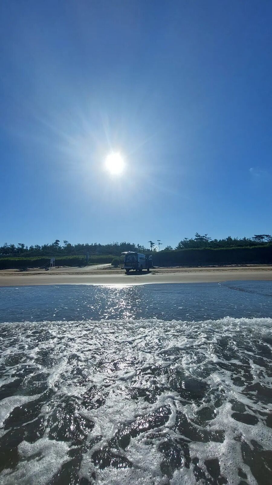 The campervan seen from the sea on the second visit, backlit by morning sun, waves sparkling in the foreground, silhouette of the van on the sand