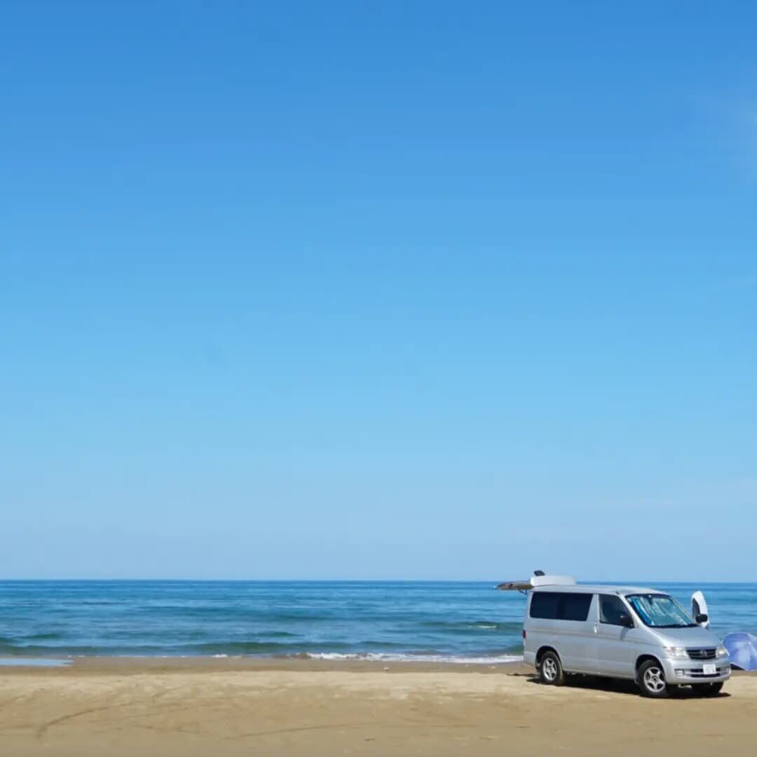 The campervan right at the waterline with the Sea of Japan and blue sky filling the frame, waves lapping at the hard-packed sand