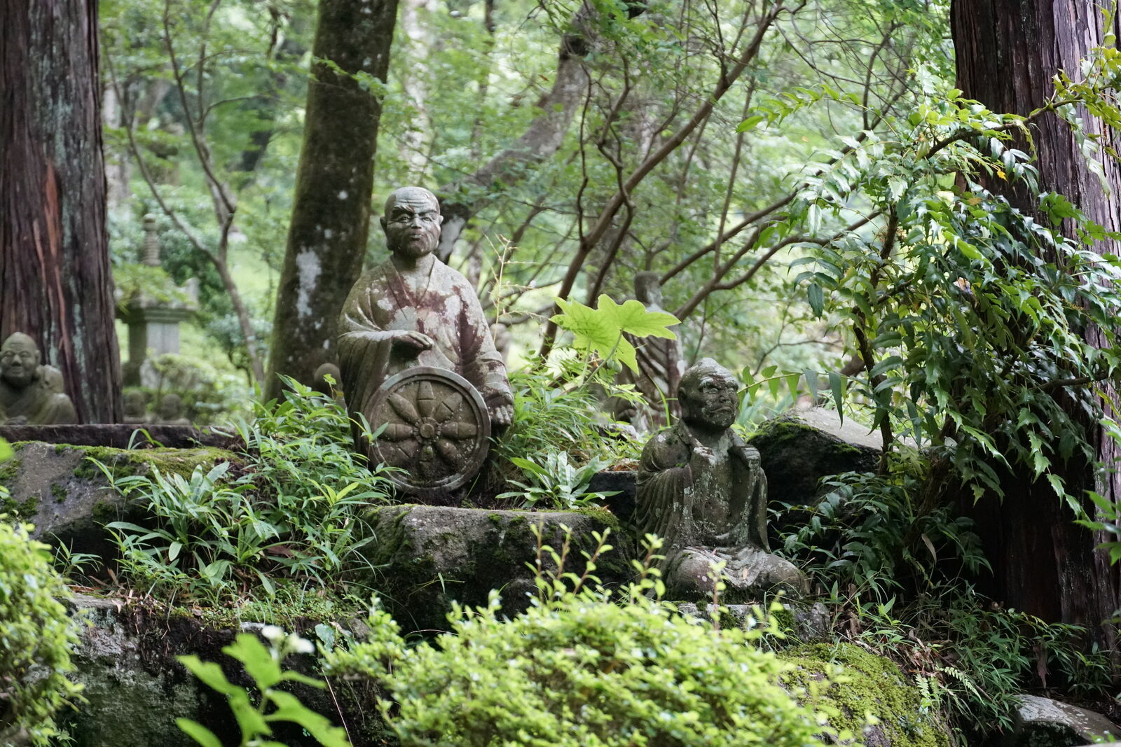 Large figure holding a dharma wheel beside a smaller figure holding up a leaf, both surrounded by lush green forest