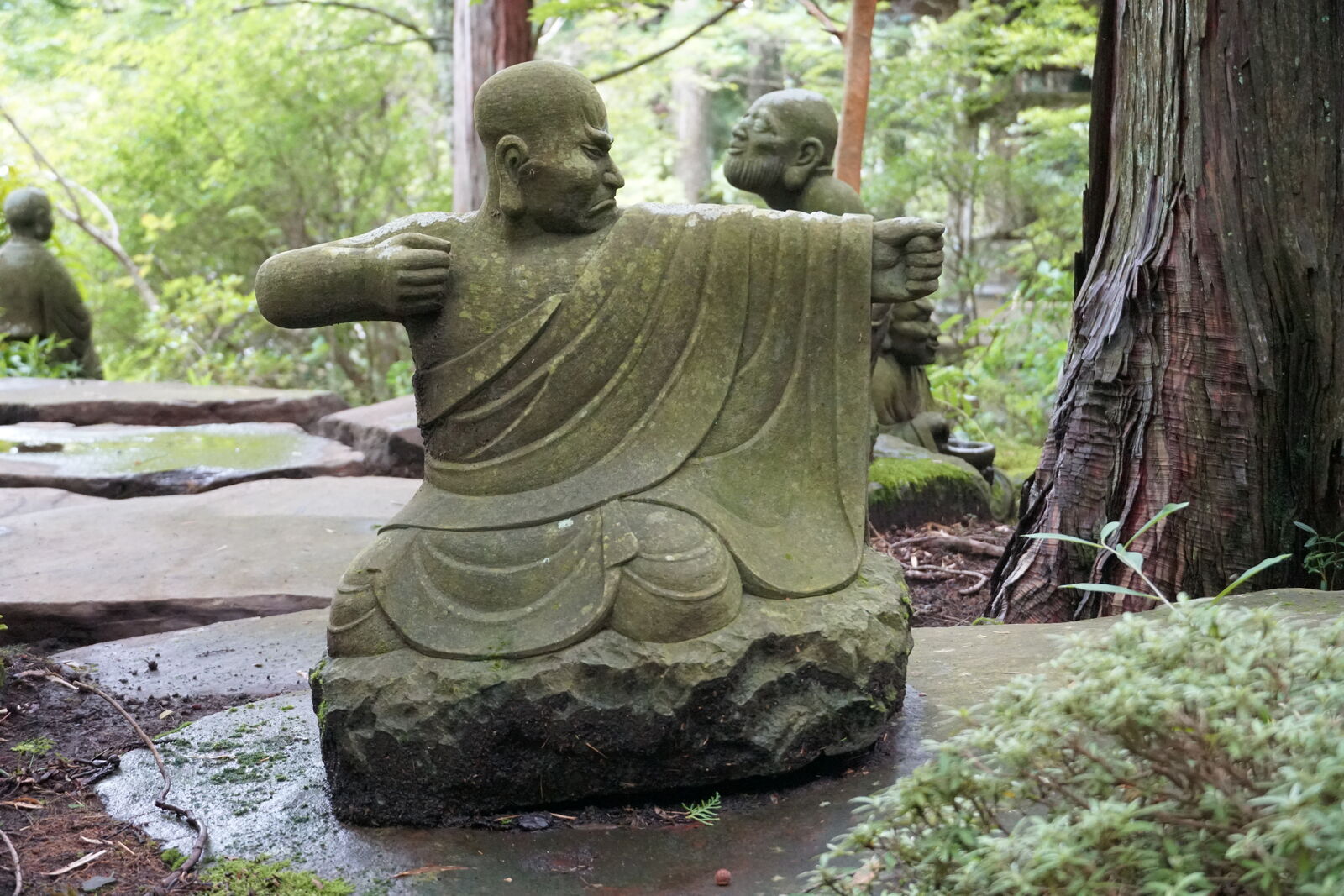 Monk figure seen from behind with arms spread wide, two smaller monks facing him, as if teaching