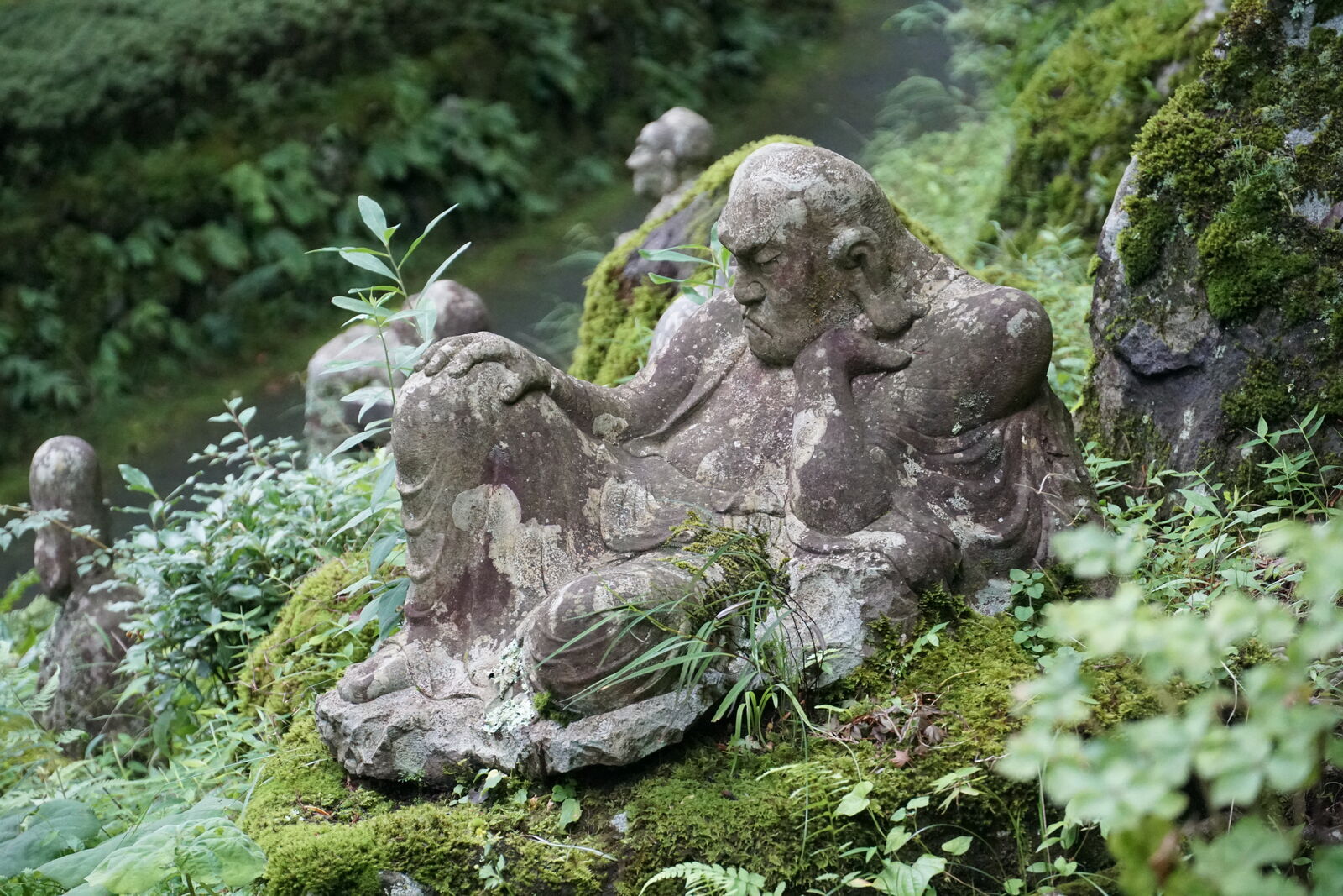 Reclining rakan figure with weathered stone and lichen, resting on a mossy rock among ferns