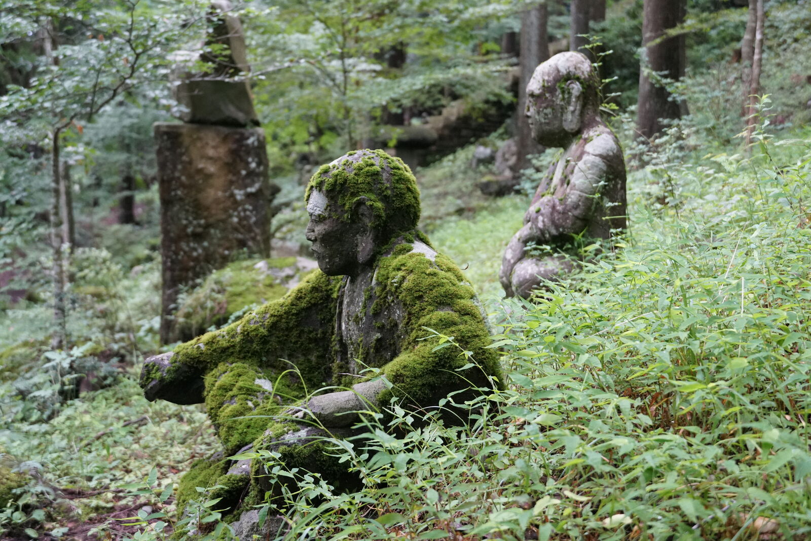 Two seated figures in conversation, one covered in moss and the other still weathered stone, forest around them