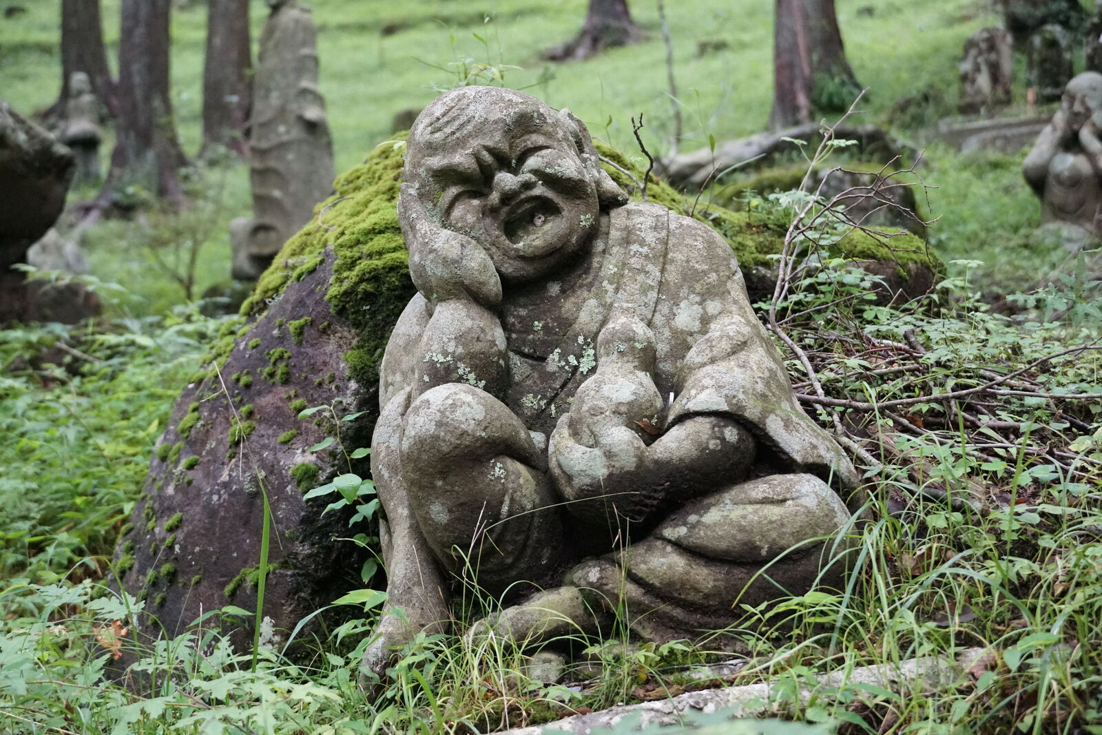Seated figure leaning against a mossy boulder, laughing with squinted eyes and open mouth, forest hillside behind