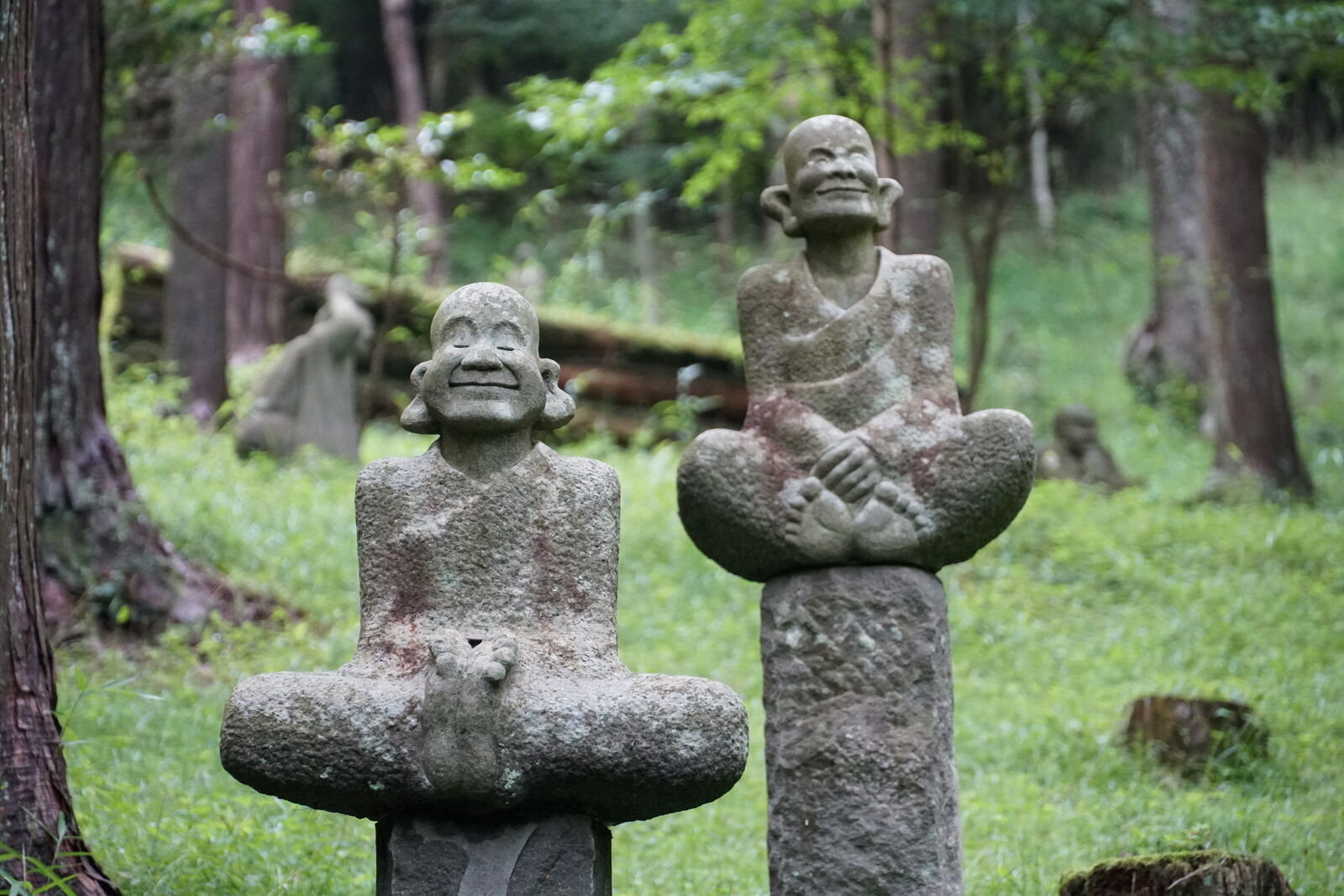 Two rakan figures on stone pillars, both looking up and laughing, green forest behind them