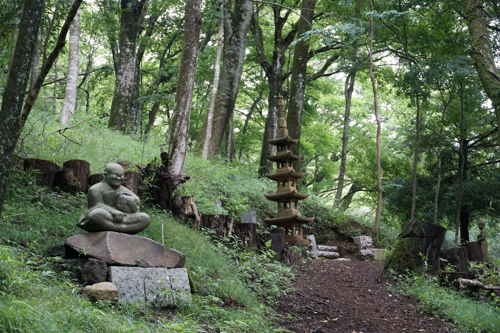 Forest path with a seated figure and a stone pagoda, the trail leading deeper into the green hillside