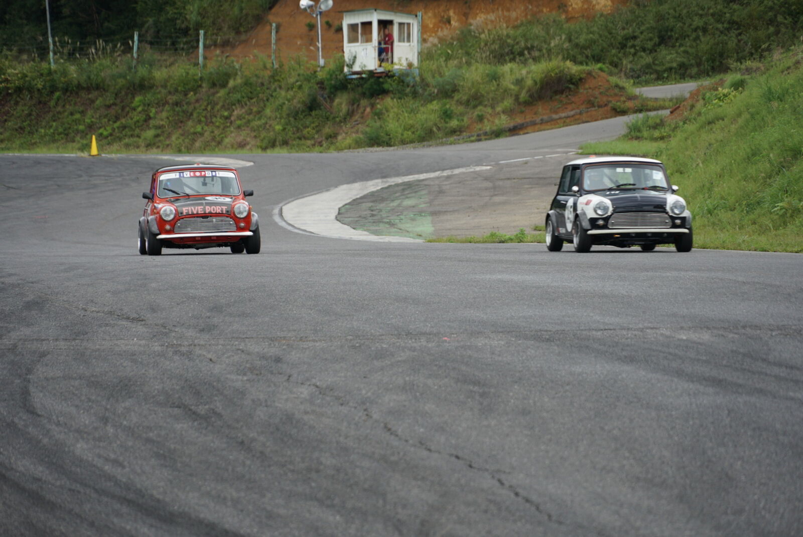 Two classic Minis racing through a banked curve on the hillside course with guard hut above