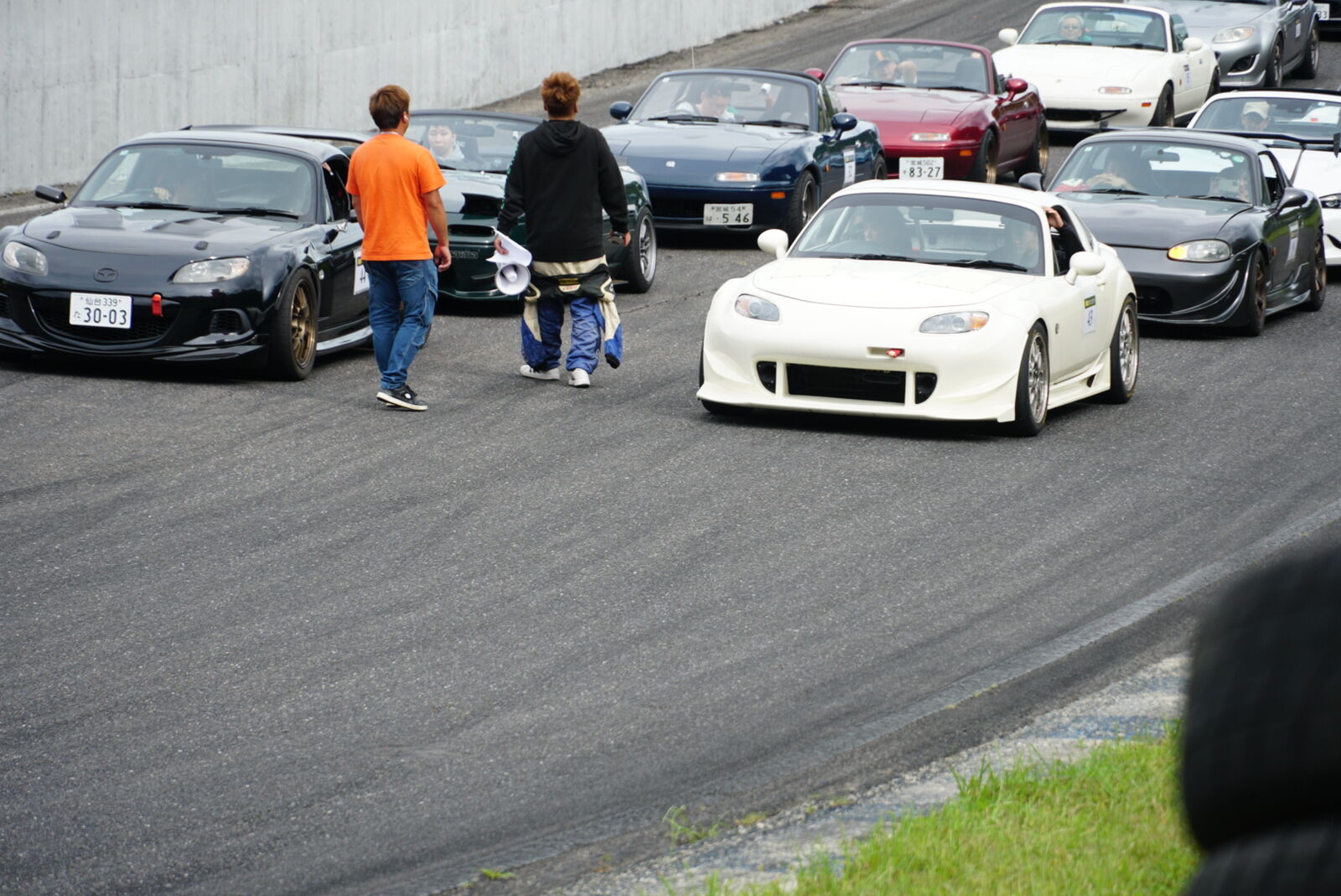 White widebody Mazda MX-5 leading a queue of sports cars with spectators and families alongside