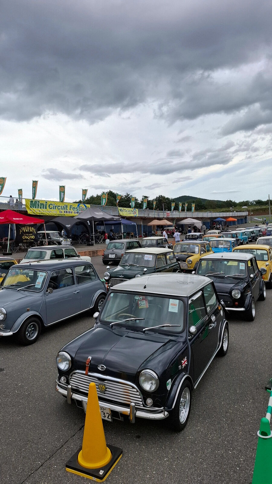 Dozens of classic Minis lined up in the parking area with dramatic cloudy sky and Mini Circuit banner