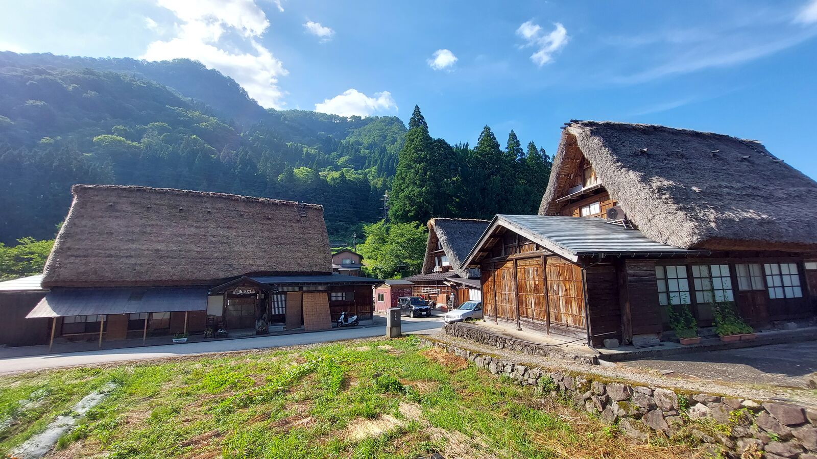 Two gassho-zukuri houses side by side with mountains and blue sky behind, stone foundation walls visible