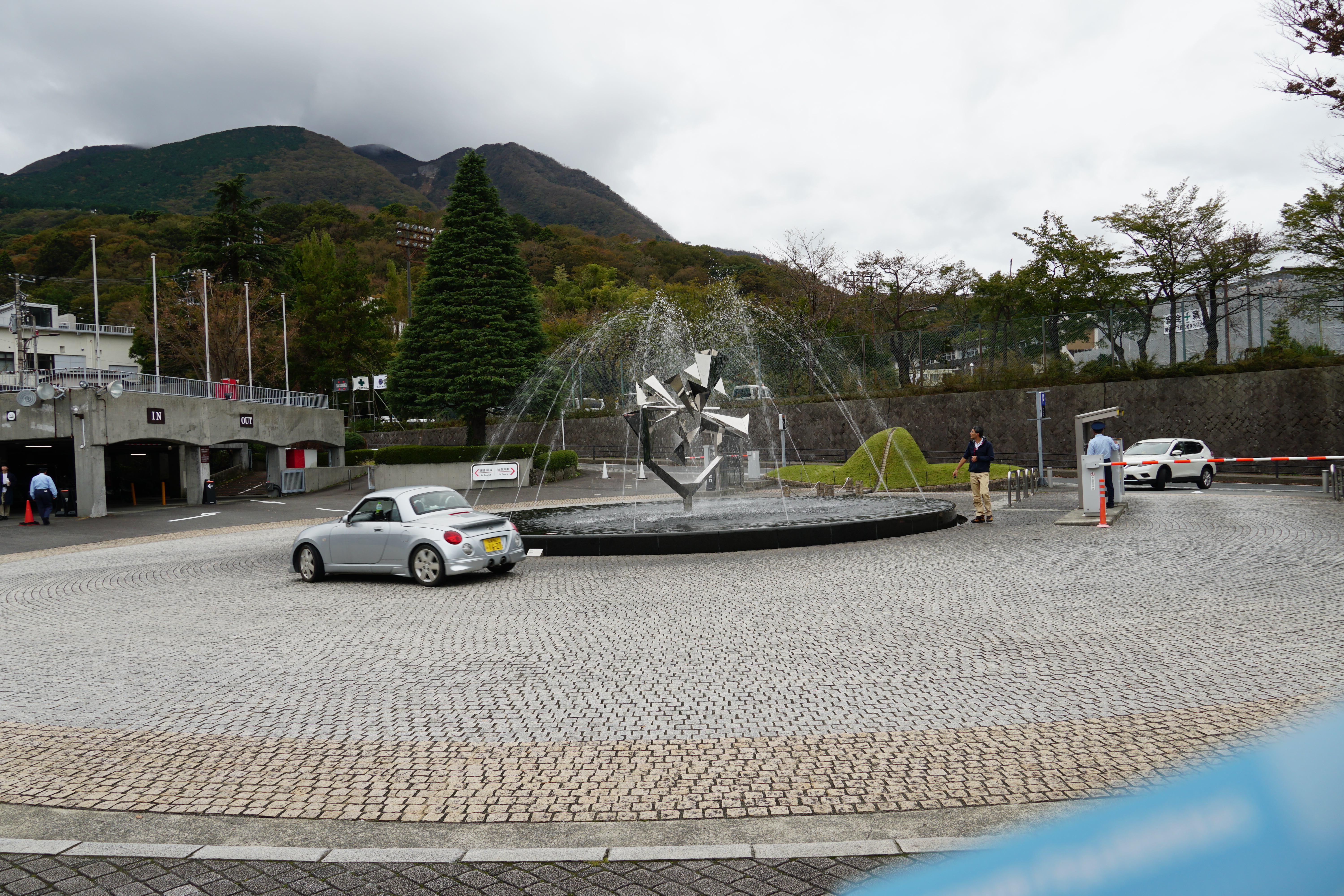Museum entrance plaza with metallic fountain sculpture, mountains rising behind the parking area