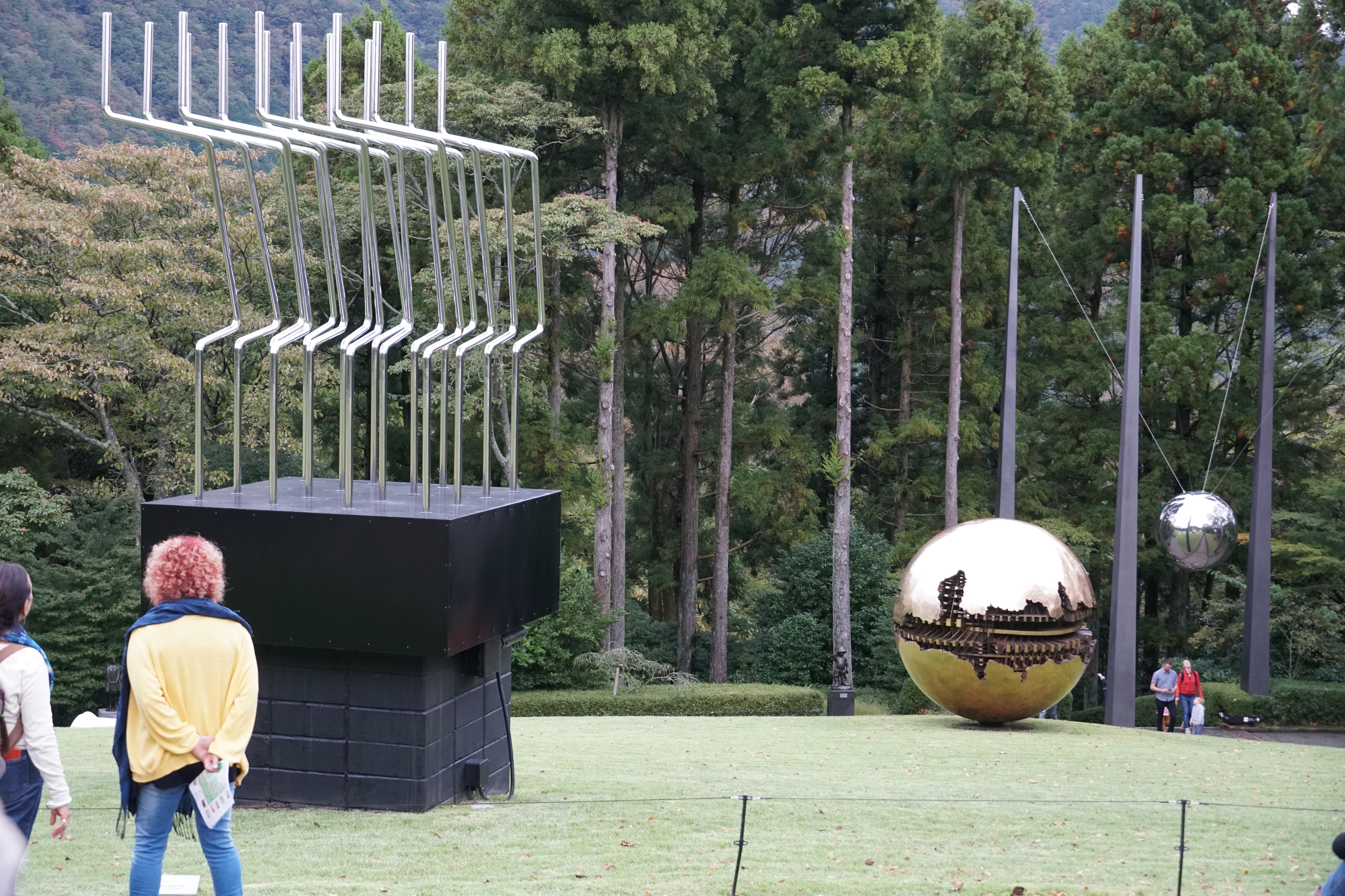 Silver pipe sculpture and golden cracked sphere on a green lawn with forest behind