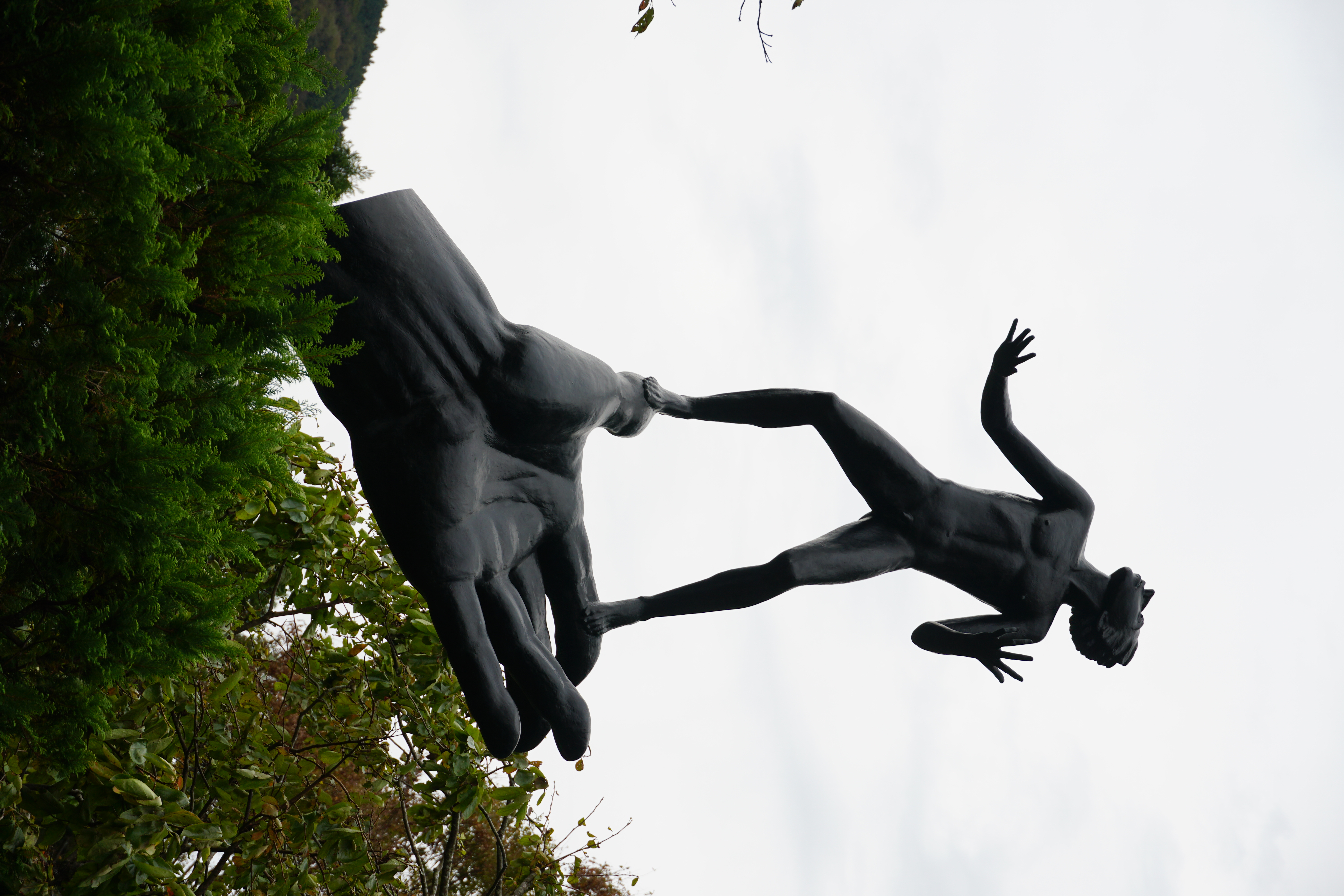 Dramatic bronze sculpture of a giant hand releasing a falling human figure, shot from below against the sky