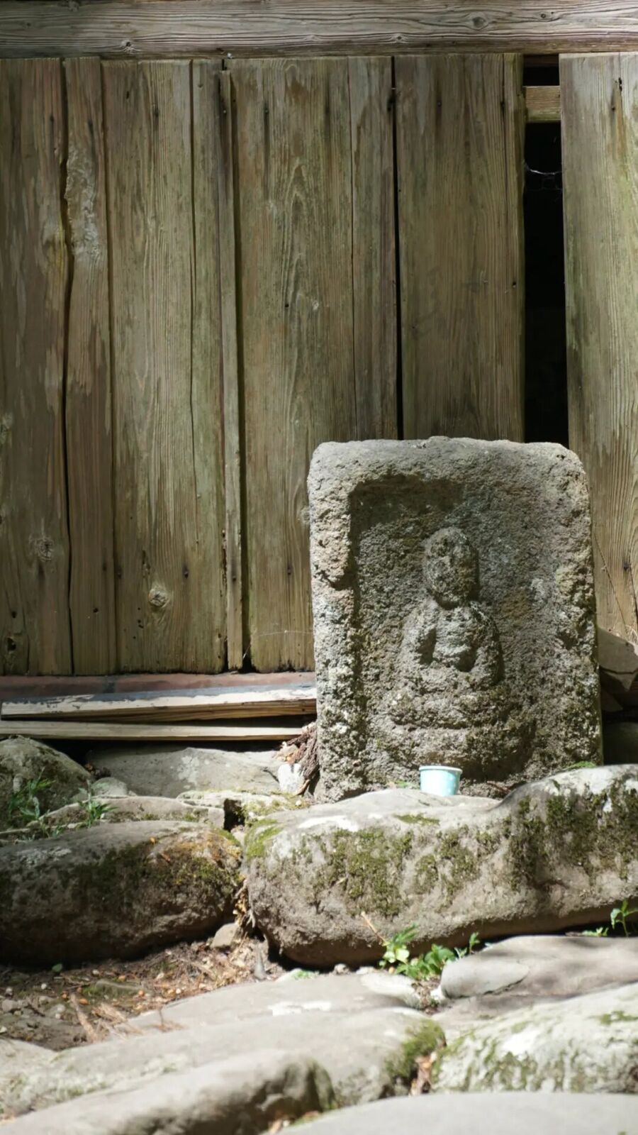 Weathered stone Buddha tablet leaning against an ancient wooden wall, a small offering cup at its base, centuries of history in the carved relief