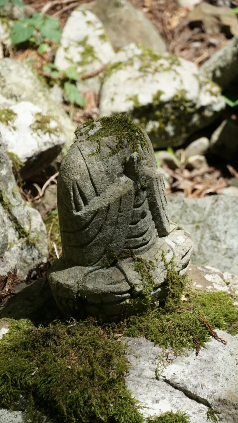Small moss-covered stone Buddha figure sitting among rocks, its features softened by centuries of moss growth, hands folded in prayer