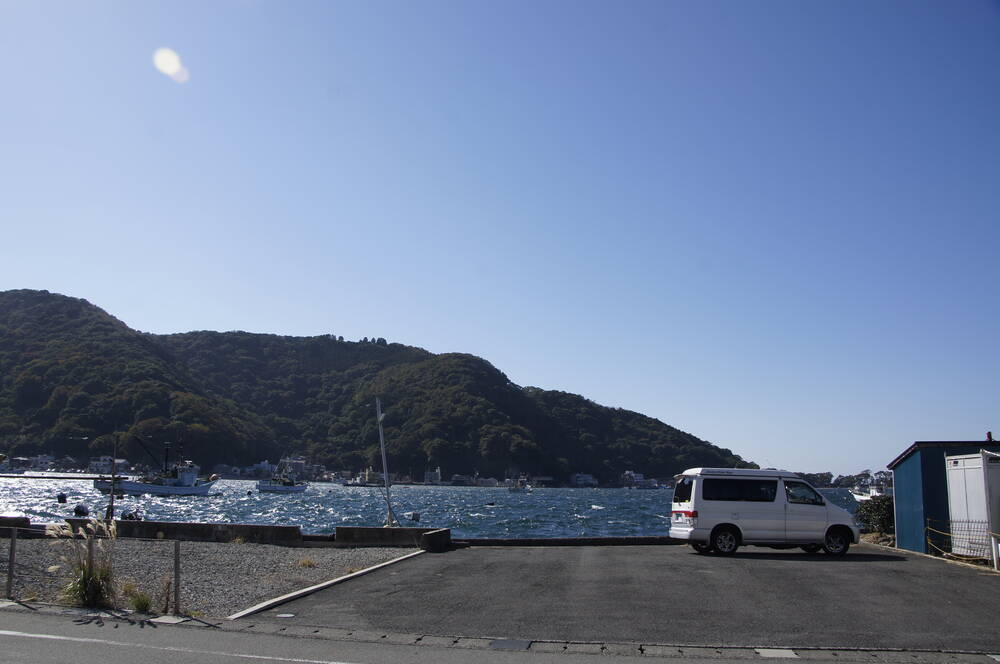 A white campervan parked at the harbour edge with the bay and fishing boats behind