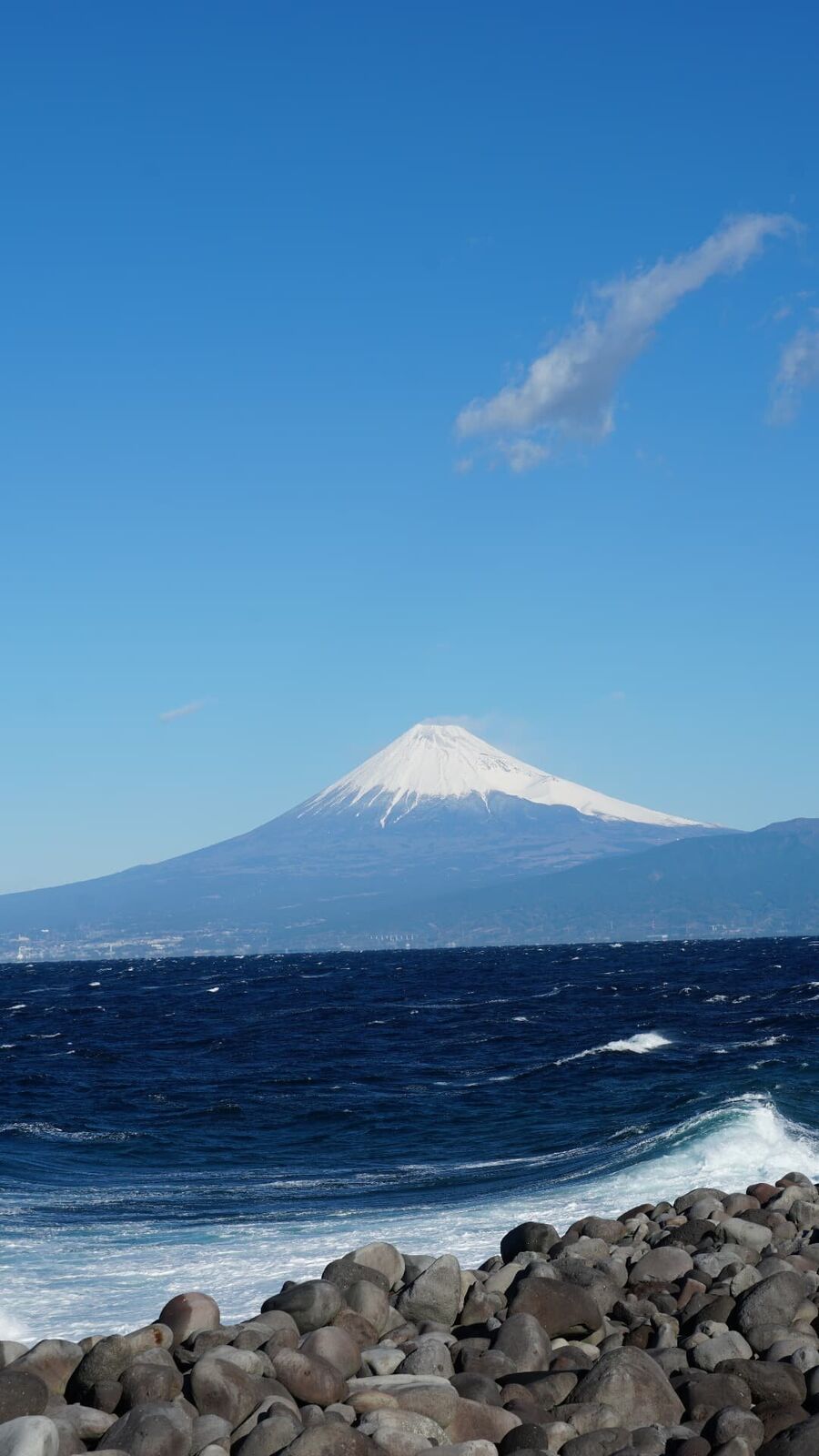 Snow-capped Mount Fuji rising above choppy Suruga Bay, waves breaking on the pebble beach in the foreground