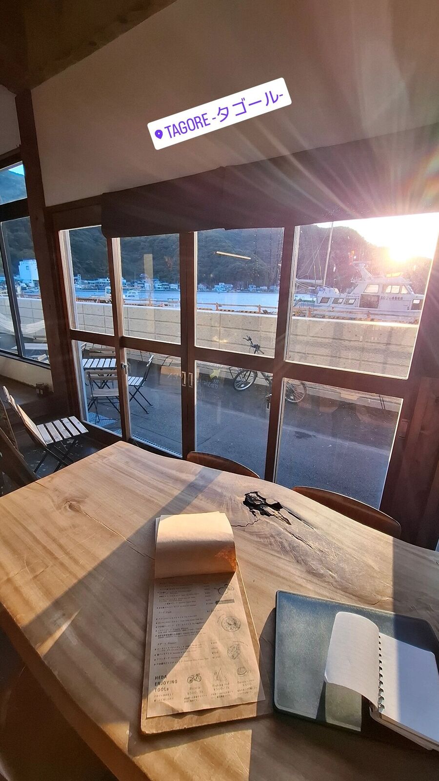 Inside Cafe Tagore at sunset, warm light streaming through the windows, the harbour and fishing boats visible outside, a menu and notebook on the live-edge wooden table