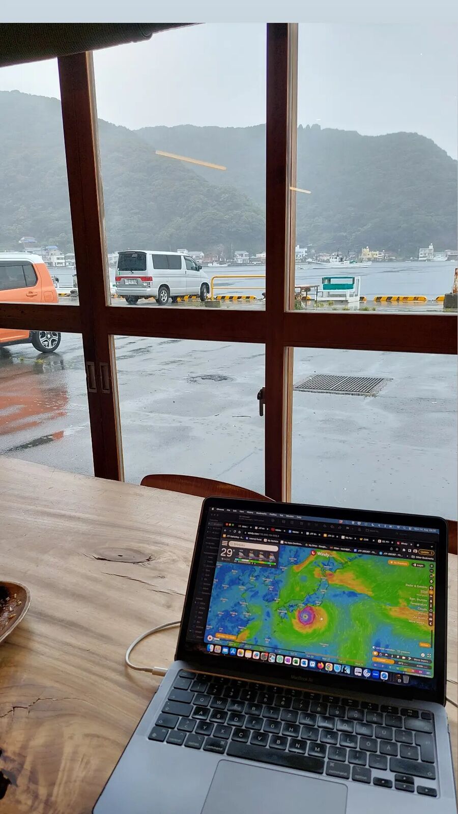 Working from Cafe Tagore during a typhoon, laptop showing the weather radar, the campervan parked outside in the rain with the harbour grey and windswept behind