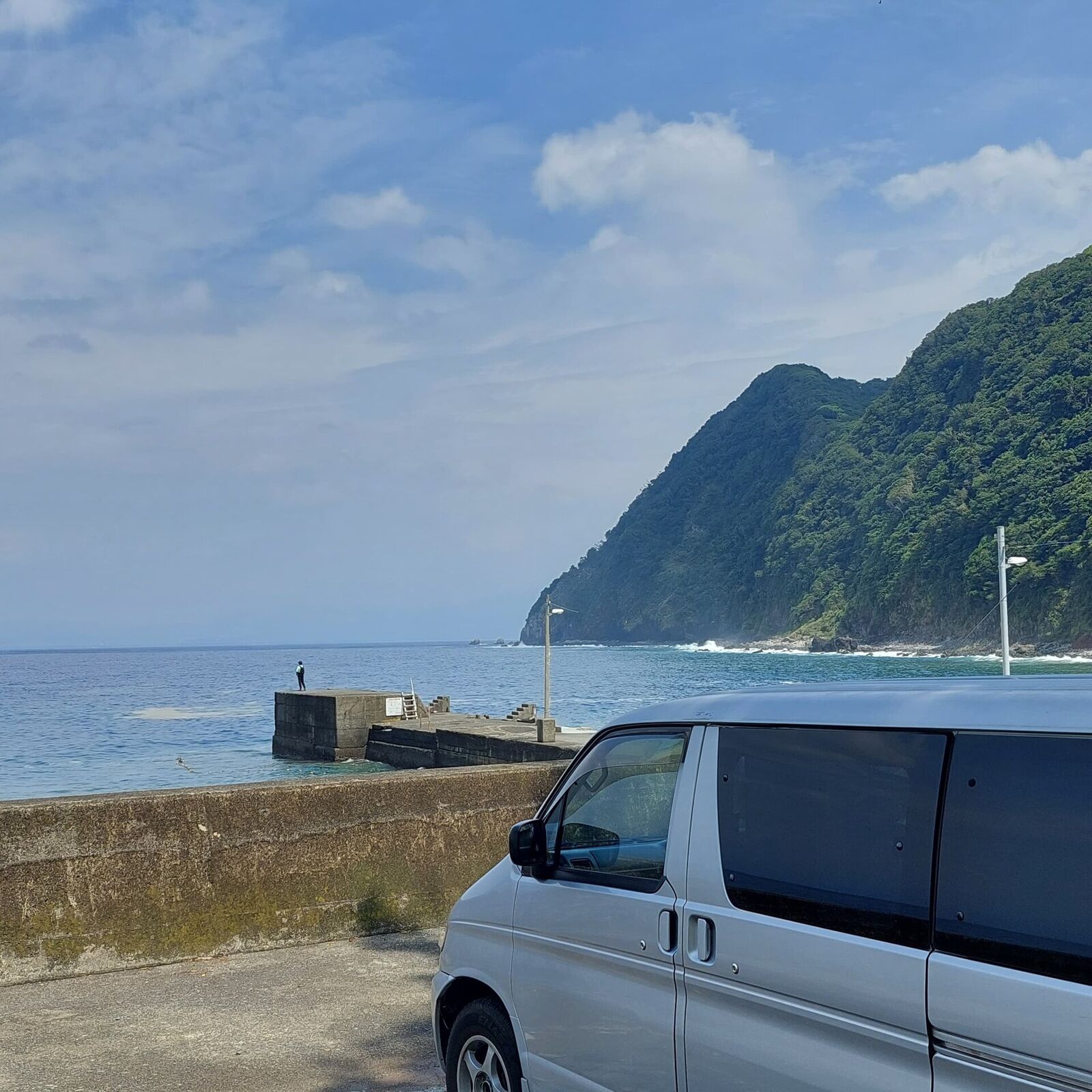 The campervan parked at a coastal harbour with a lone fisherman on the pier, dramatic cliff and deep blue sea stretching to the horizon