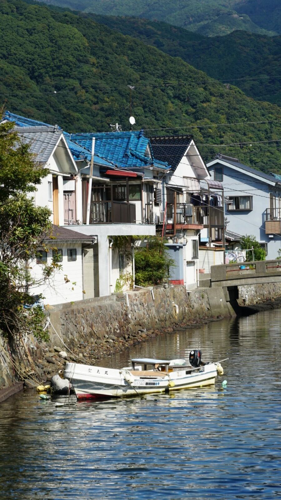 Heda village waterfront with traditional houses, blue-tiled roofs, a small boat in the canal, and green mountains rising behind