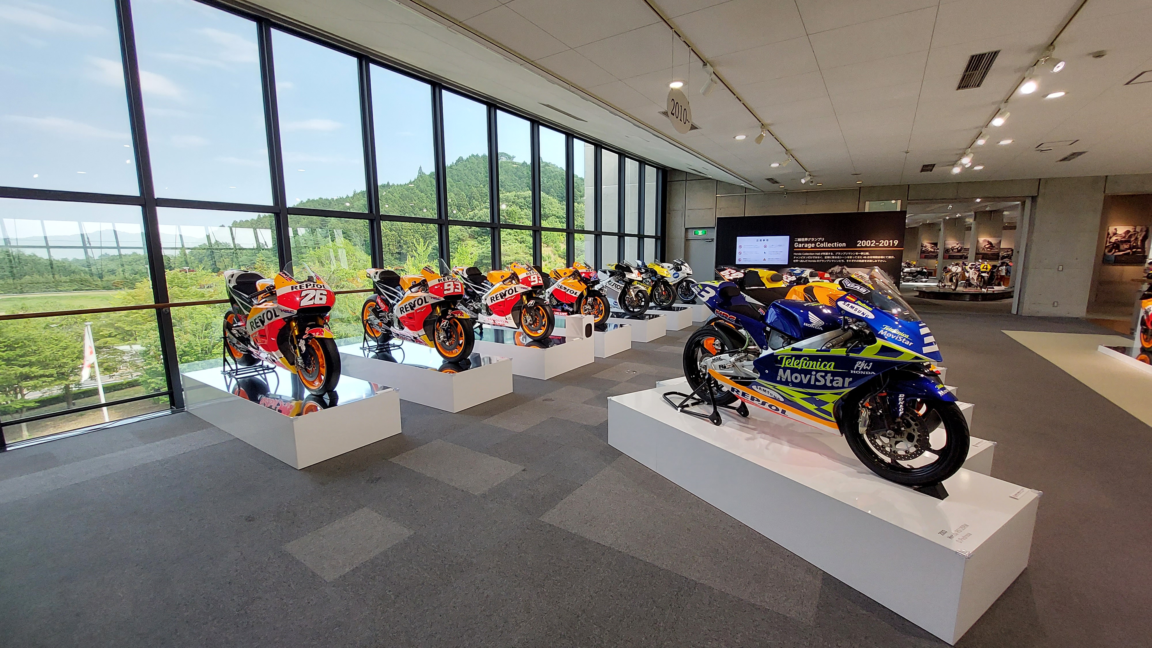 MotoGP bikes lined up against the floor-to-ceiling windows, Movistar and Repsol liveries with green forest behind the glass