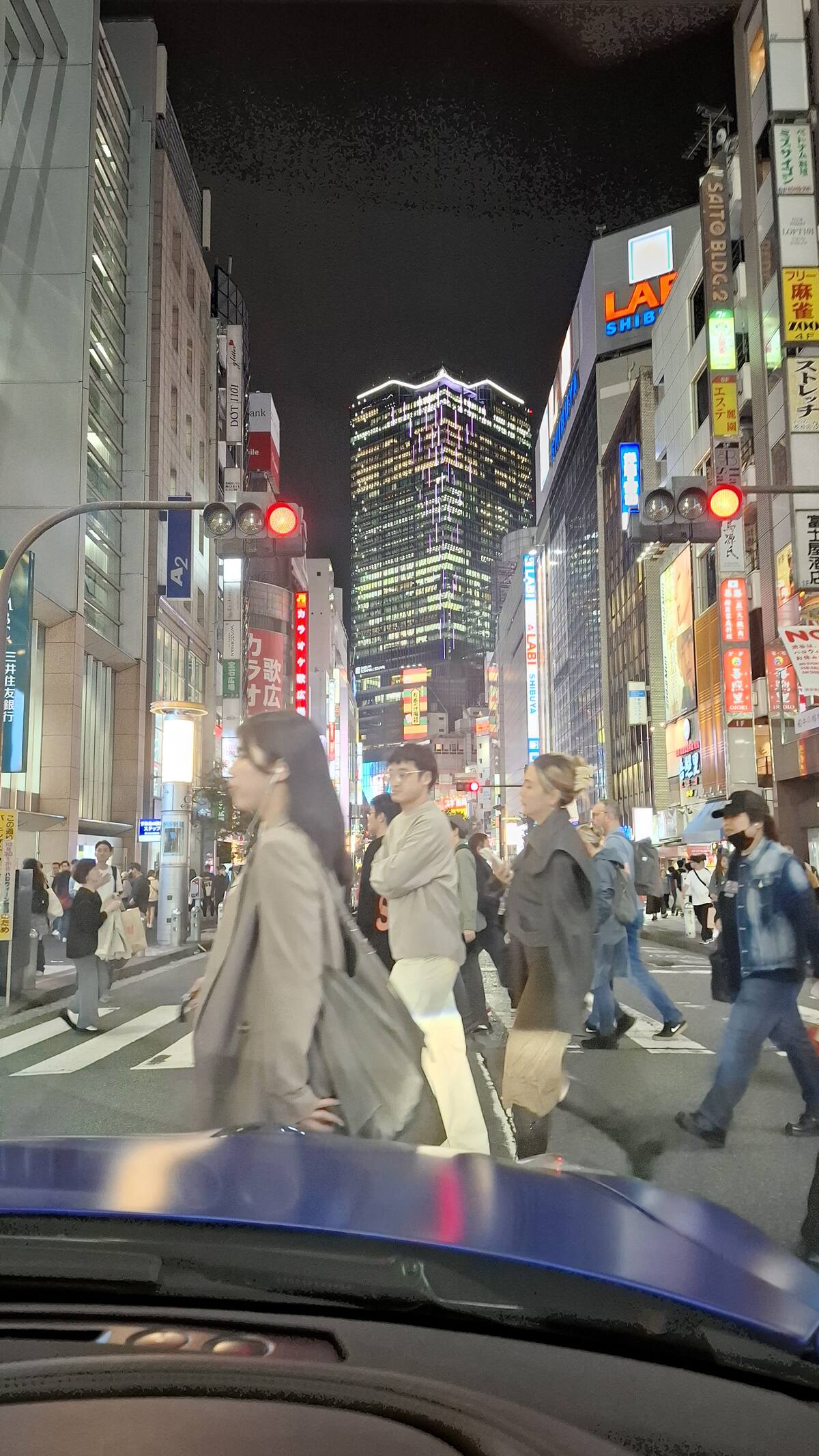View from the GT-R cockpit driving through Shibuya at night with neon signs and pedestrians