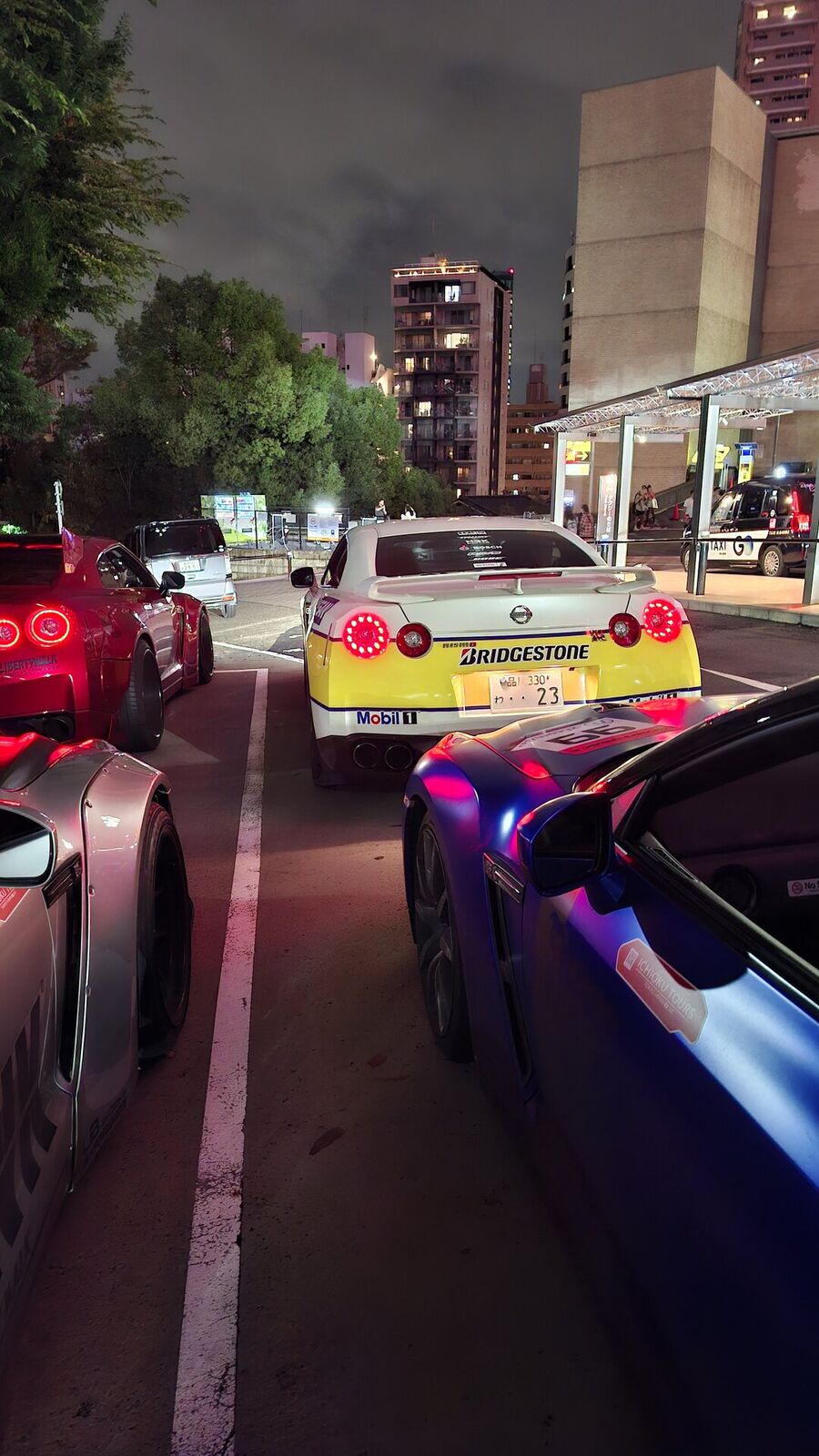 The convoy from behind at a stop: yellow Bridgestone-livery GT-R #23, red GT-R, and blue GT-R #99, their taillights reflecting on wet asphalt