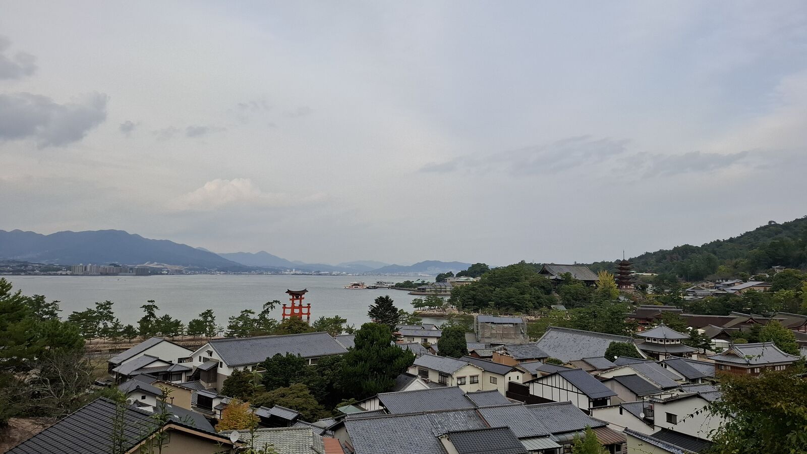 View from the hillside above Miyajima town: traditional tiled rooftops stretching toward the water, the floating torii and five-story pagoda visible, the Inland Sea and mountains in the distance