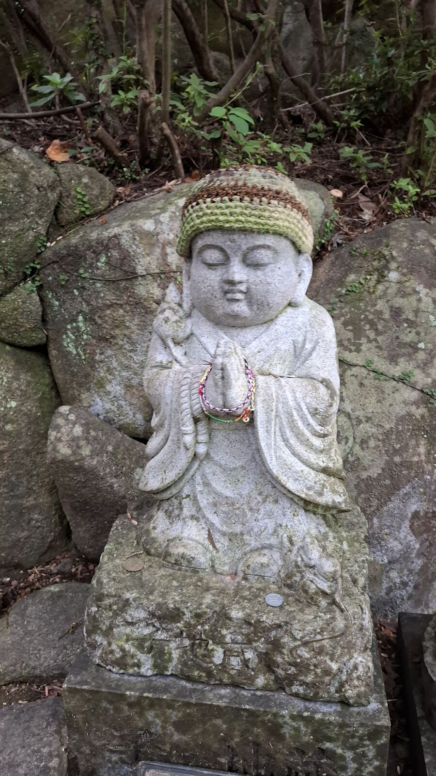 A serene jizo statue with a hand-knitted green cap, eyes peacefully closed, the detailed stone carving showing flowing robes and a gentle smile
