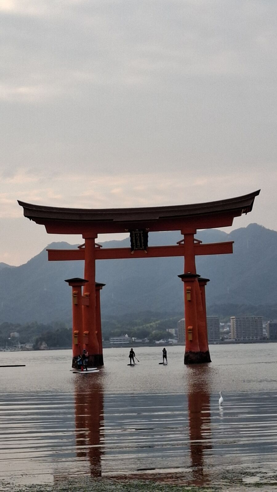 The torii gate at low tide with people walking on the exposed seabed toward it, shallow pools reflecting the sky, the gate's full wooden structure visible from base to top