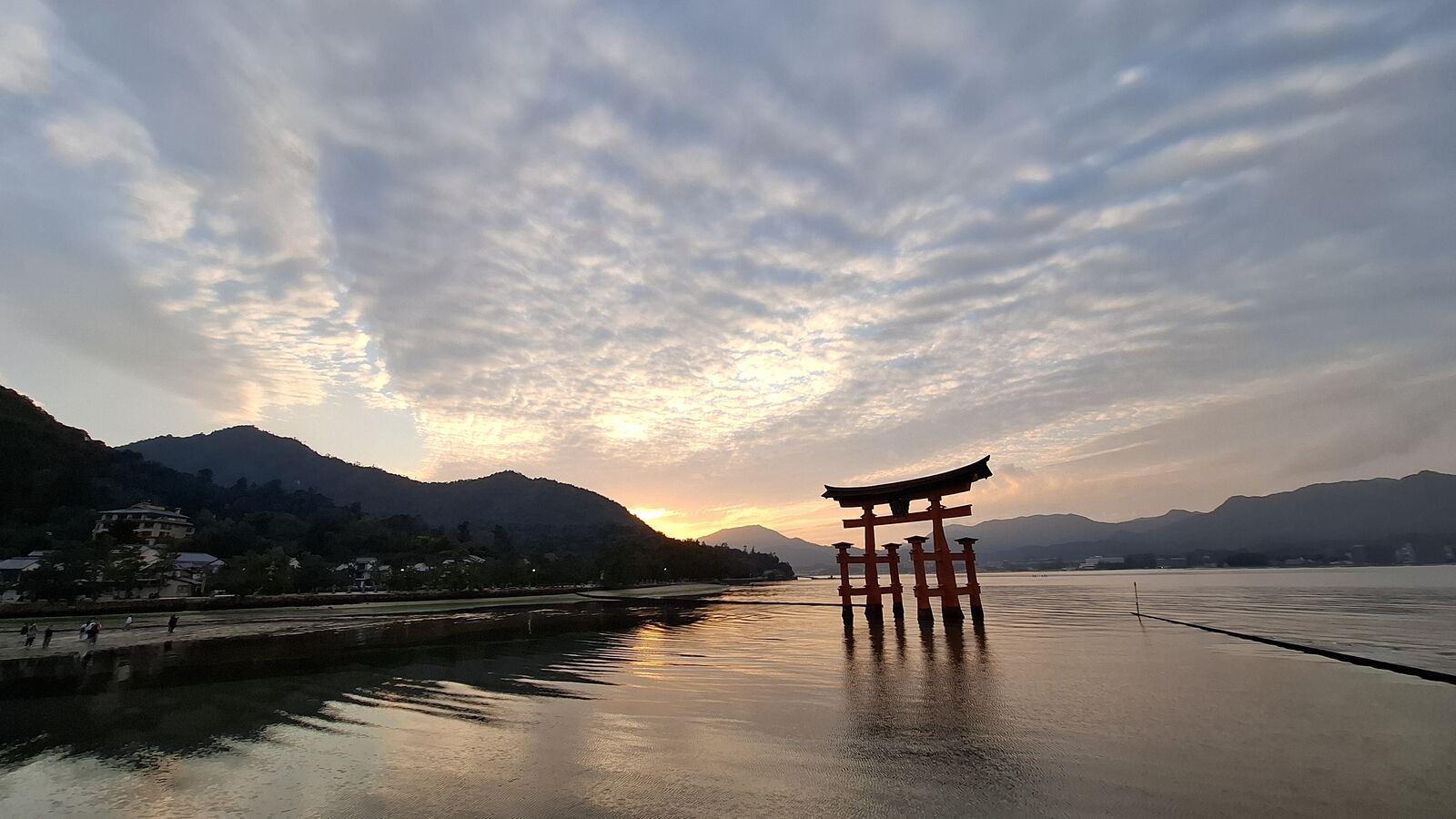 The floating torii of Itsukushima Shrine silhouetted against a golden sunset sky, Miyajima's forested mountain rising behind it, golden reflections shimmering in the shallow water