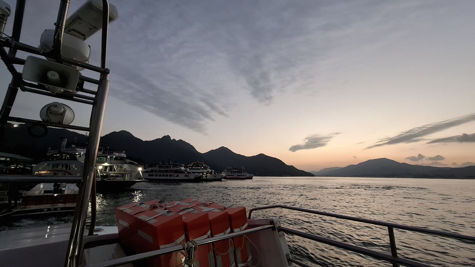 On the return ferry at dusk, life jacket containers visible in the foreground, ferries and the island's mountains behind, the sky turning to evening