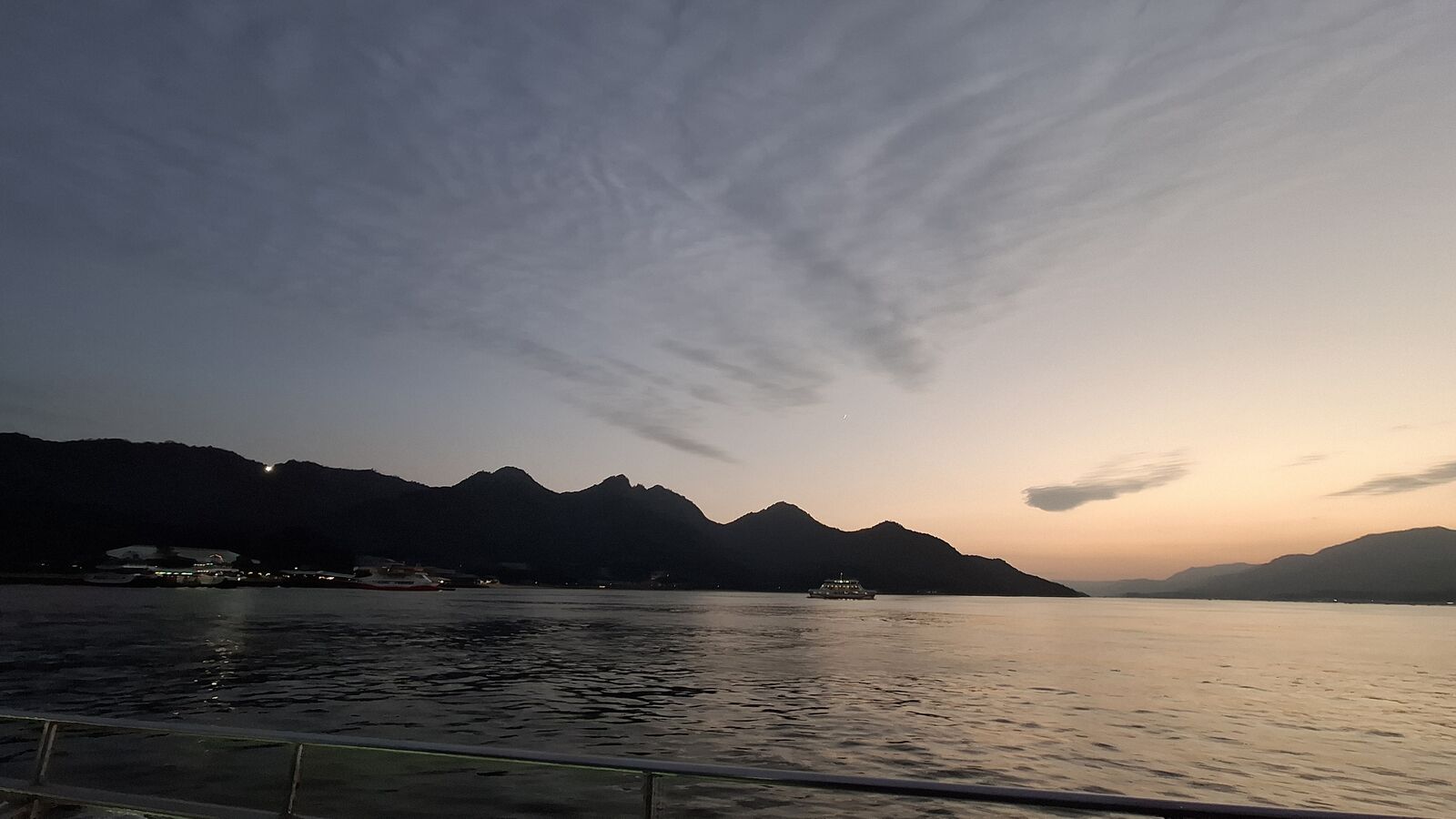Miyajima's mountain silhouette at dusk seen from the return ferry, a ferry crossing in the distance, crescent moon visible in the evening sky, the day ending as quietly as it began