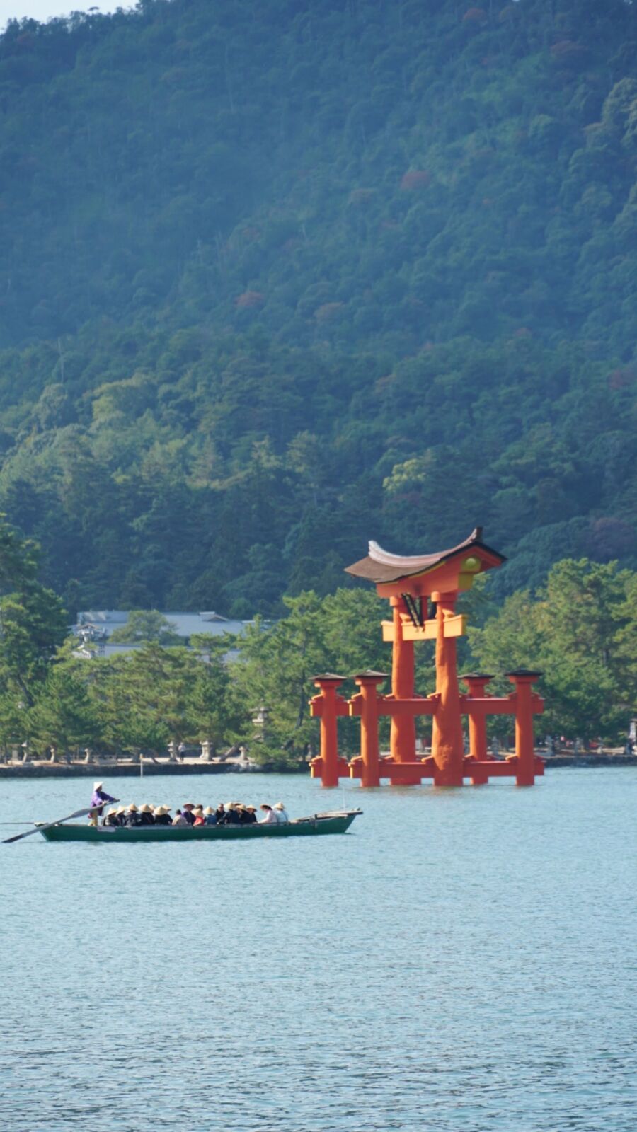 A traditional rowing boat passing in front of the floating torii at high tide, tourists wearing straw hats while the boatman stands at the stern, Miyajima's forested mountain filling the background