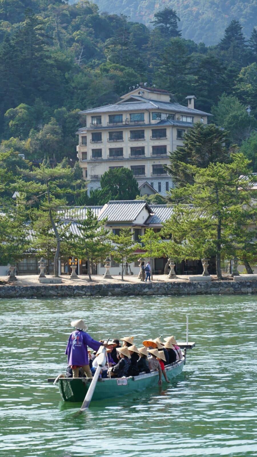 Close-up of the tour boat: tourists in traditional straw hats being rowed past the island's shore by a boatman in purple traditional dress, a ryokan visible on the hillside behind
