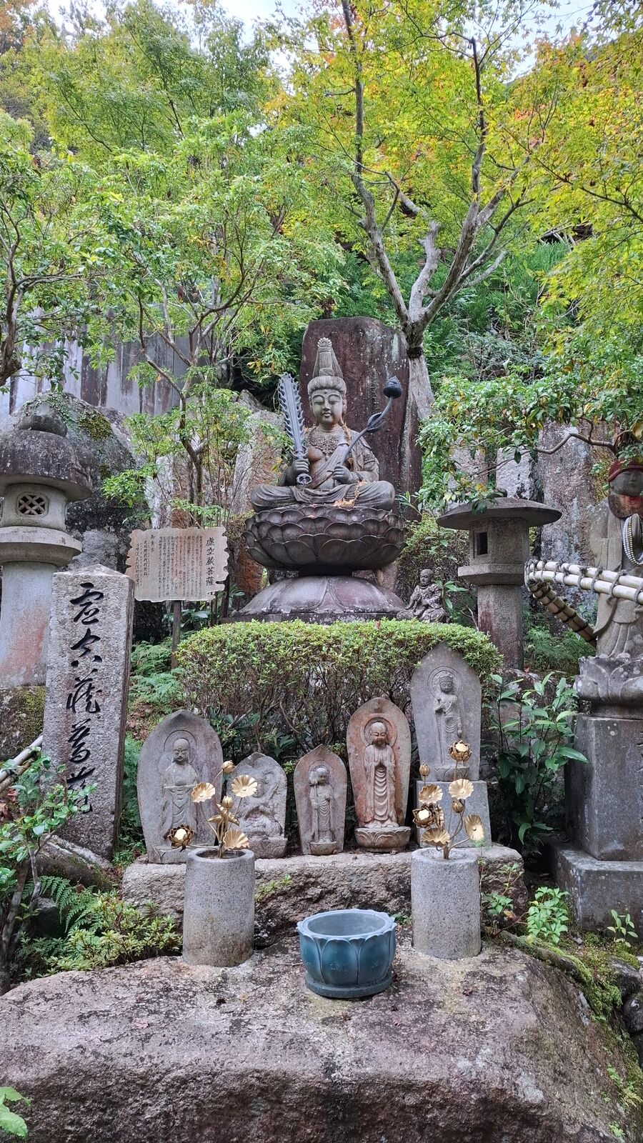Buddhist statue arrangement at Daisho-in: a seated Kannon figure on a mossy pedestal surrounded by smaller stone figures with golden ornaments, green maple leaves framing the scene