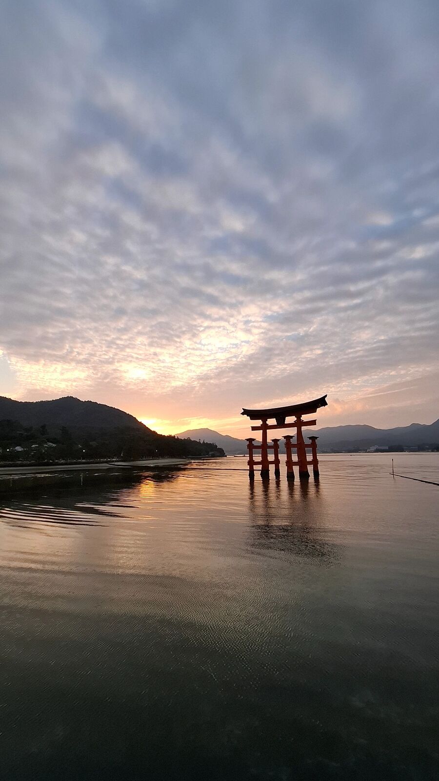 Vertical view of the torii at sunset, the gate standing in shallow golden water with dramatic cloud formations spreading across the sky above