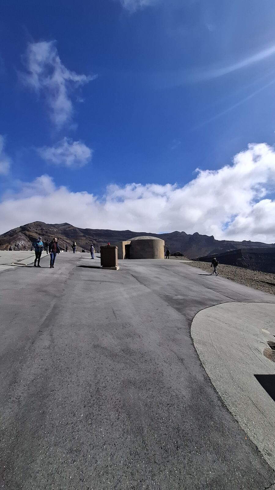 The volcanic moonscape around the crater with paved paths, evacuation bunkers, and a handful of visitors walking in the distance under a vast sky