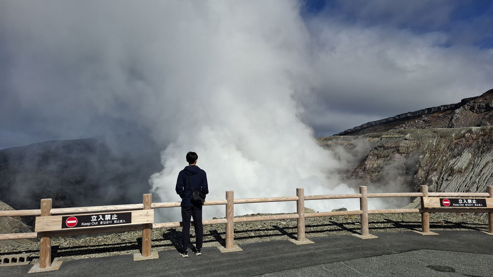 Kevin standing at the railing overlooking the crater, a wall of volcanic steam rising behind him, Keep Out sign visible on the fence
