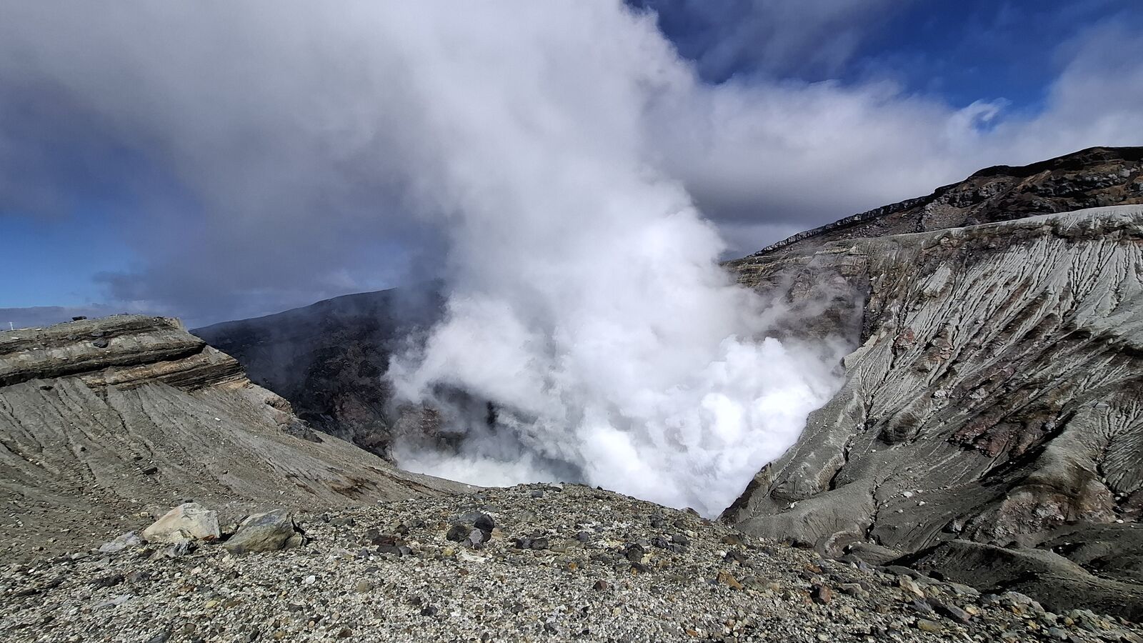 Looking into the active crater of Nakadake, massive steam column rising from the depths, layered geological formations of grey, brown and rust surrounding the vent