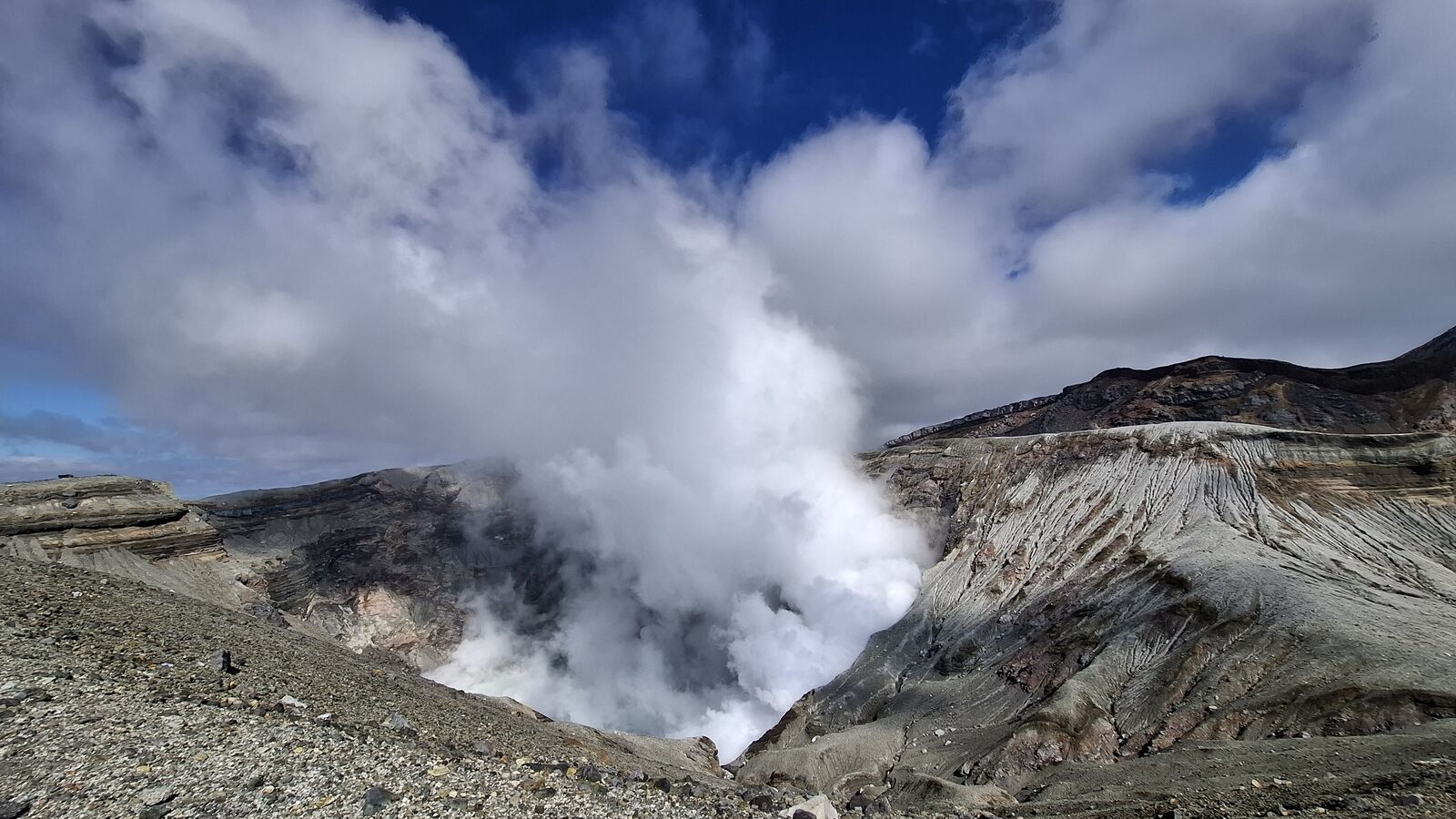 The crater from a different angle, steam billowing upward against blue sky, striped crater walls showing layers of volcanic deposits