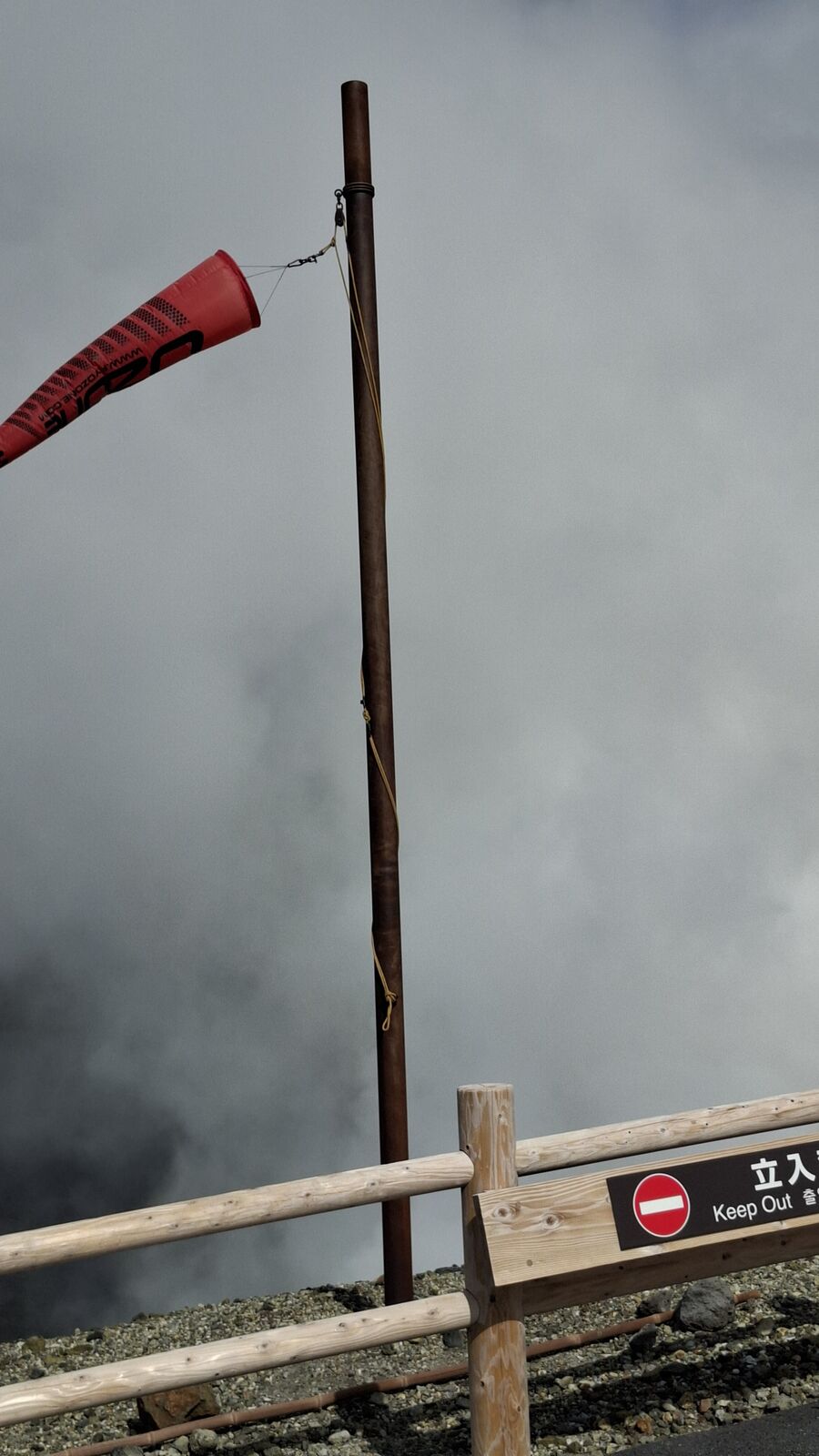 Red windsock and Keep Out sign at the crater's edge, volcanic gas drifting past in the background, a tool for measuring wind direction and gas flow
