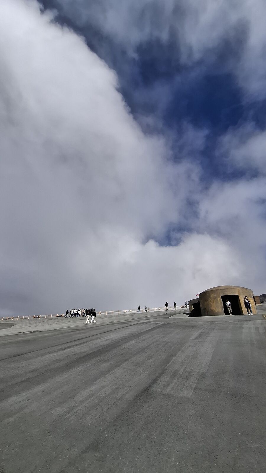 Massive volcanic steam cloud filling the sky above the crater, a concrete evacuation bunker in the foreground with tiny visitors walking the path in the distance, showing the enormous scale of the volcano against human infrastructure