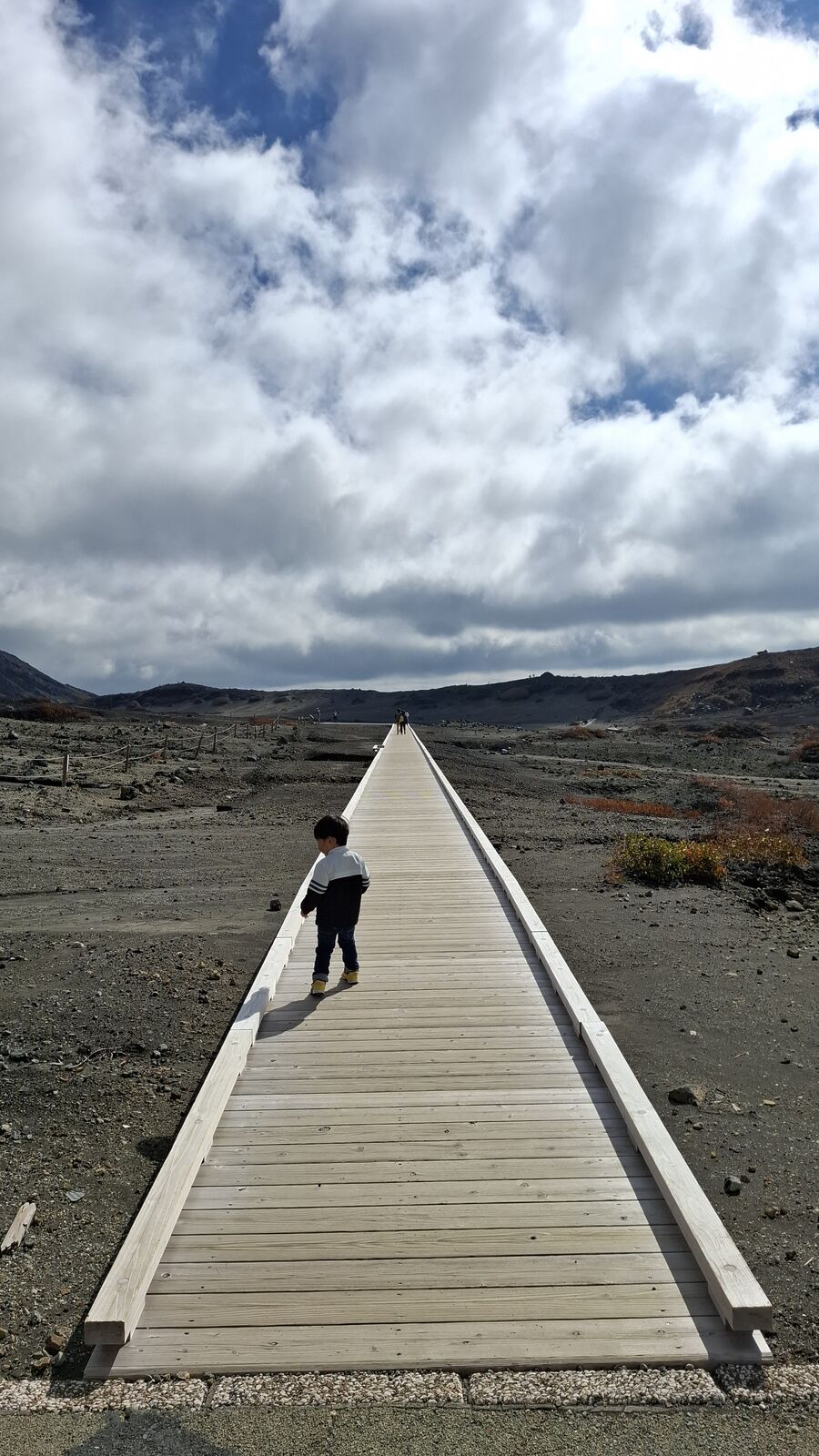 A small figure walking alone on a wooden boardwalk across Mount Aso's volcanic desert